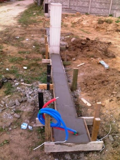 Wylewka fundamentu pod ogrodzenie betonowe z widocznymi szalunkami, zbrojeniami i rurami instalacyjnymi w ziemi.