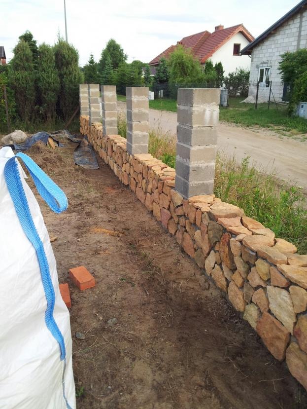 Wznoszenie kamiennego muru ogrodzeniowego z betonowymi słupkami w trakcie budowy, widoczny worek z materiałem i cegły na ziemi.