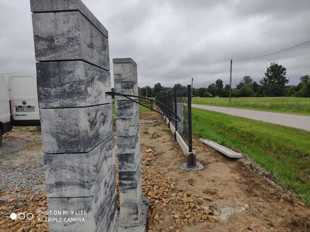 Wznoszenie ogrodzenia: betonowe słupki modułowe w trakcie montażu, widoczna czarna rura instalacyjna i fragment ogrodzenia panelowego z podmurówką prefabrykowaną, w tle krajobraz wiejski.