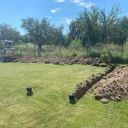 Żółta mini koparka CAT obok wykopanego rowu w trawiastym ogrodzie, widoczne czarne studzienki kanalizacyjne, w tle drzewa i ogrodzenie.