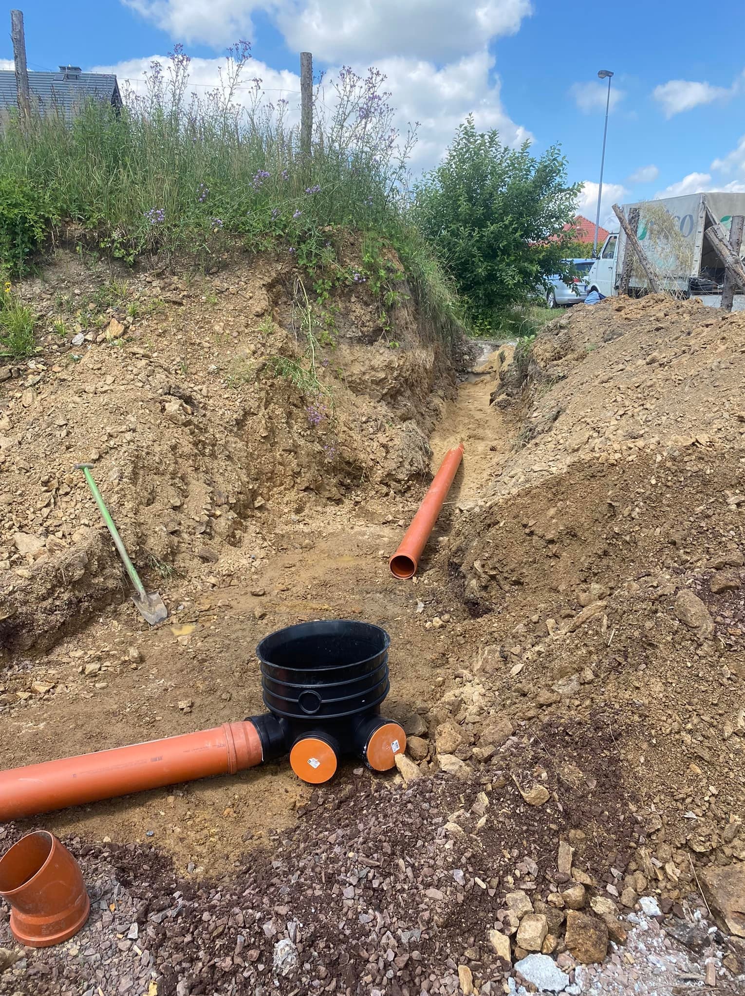 Wykonany wykop pod instalację, widoczne rury kanalizacyjne z tworzywa sztucznego, czarna studzienka rewizyjna oraz łopata wbita w ziemię. W tle krajobraz z zabudowaniami i zielenią.