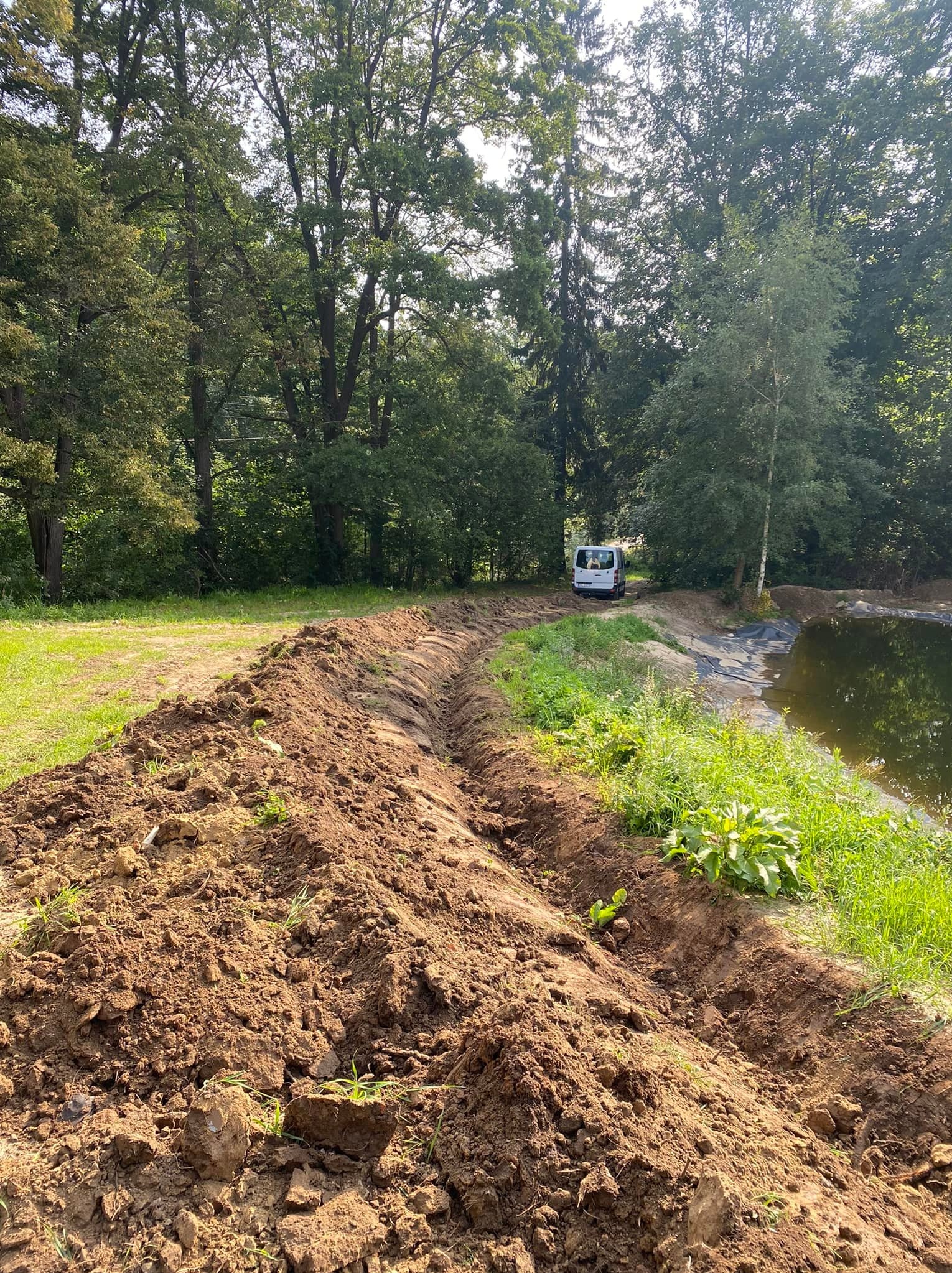 Wykopany rów w ziemi wzdłuż stawu, z widocznym białym vanem w tle na skraju lasu.