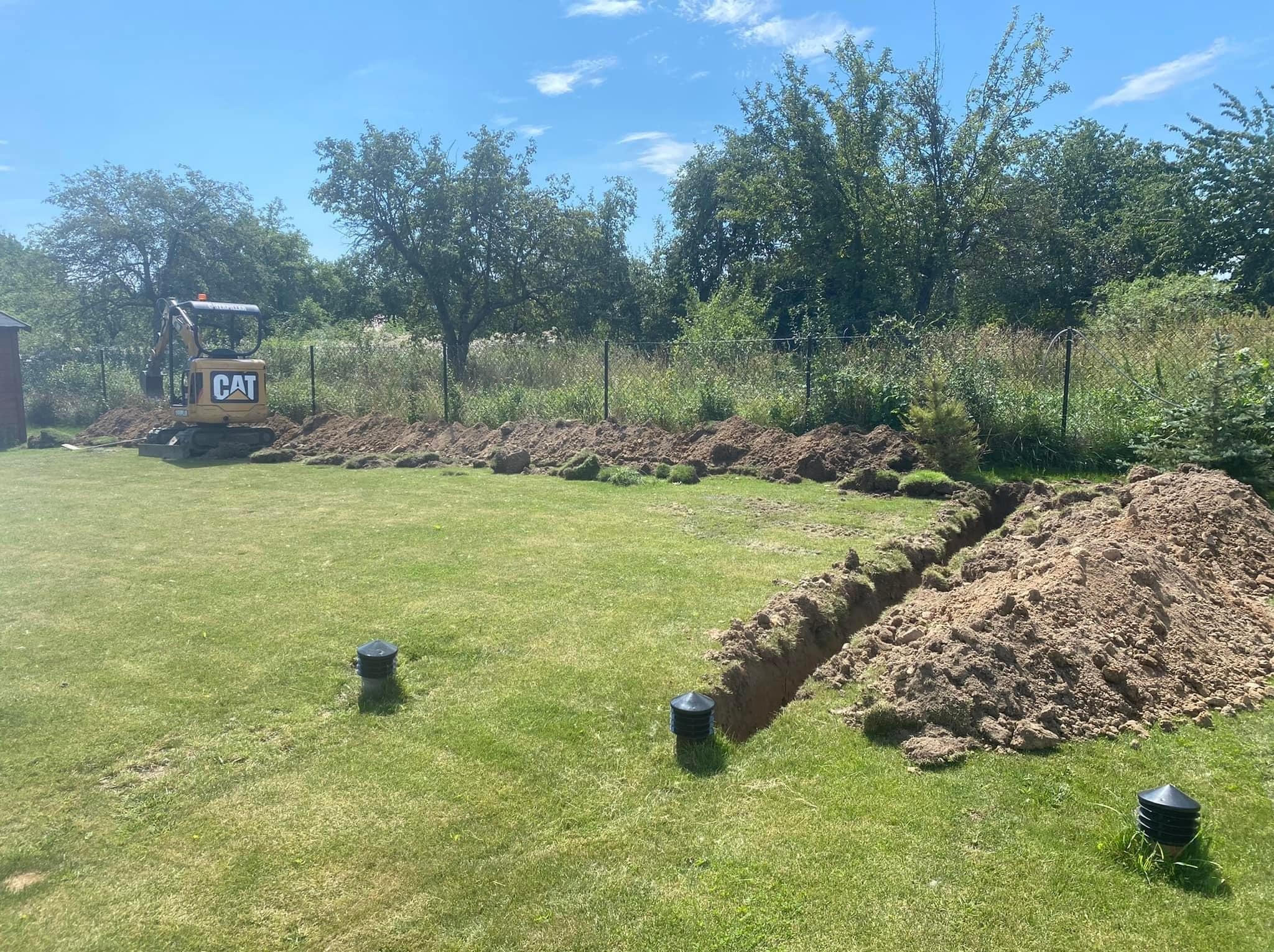 Żółta mini koparka CAT obok wykopanego rowu w trawiastym ogrodzie, widoczne czarne studzienki kanalizacyjne, w tle drzewa i ogrodzenie.
