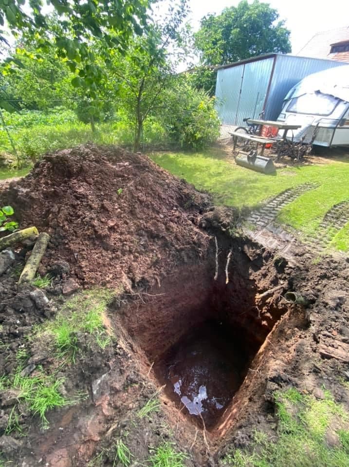 Wykopany dół w ziemi z wodą na dnie, obok sterta ziemi i zielona trawa w tle.