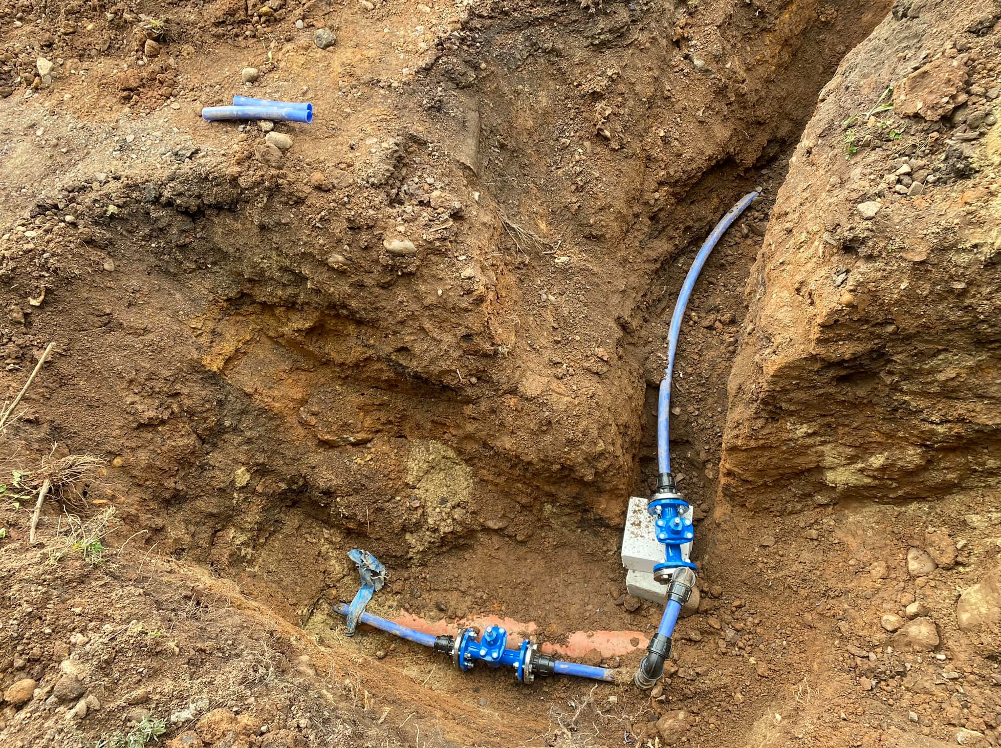 Wykonany wykop w ziemi z widocznymi niebieskimi rurami i złączkami instalacji wodociągowej, częściowo ułożonymi na cegłach.