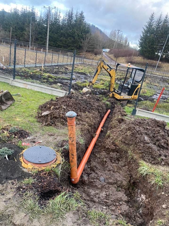 Pomarańczowe rury kanalizacyjne ułożone w wykopie, widoczny mały żółty koparka Caterpillar, studzienka kanalizacyjna z szarą pokrywą, ziemia i trawa po deszczu, w tle ogrodzenie i drzewa.