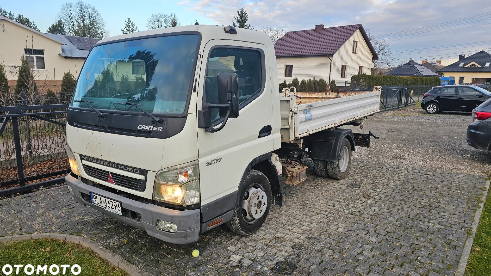 Biały samochód ciężarowy Mitsubishi Fuso Canter z otwartą skrzynią ładunkową na brukowanej powierzchni, z widocznymi domami jednorodzinnymi w tle.