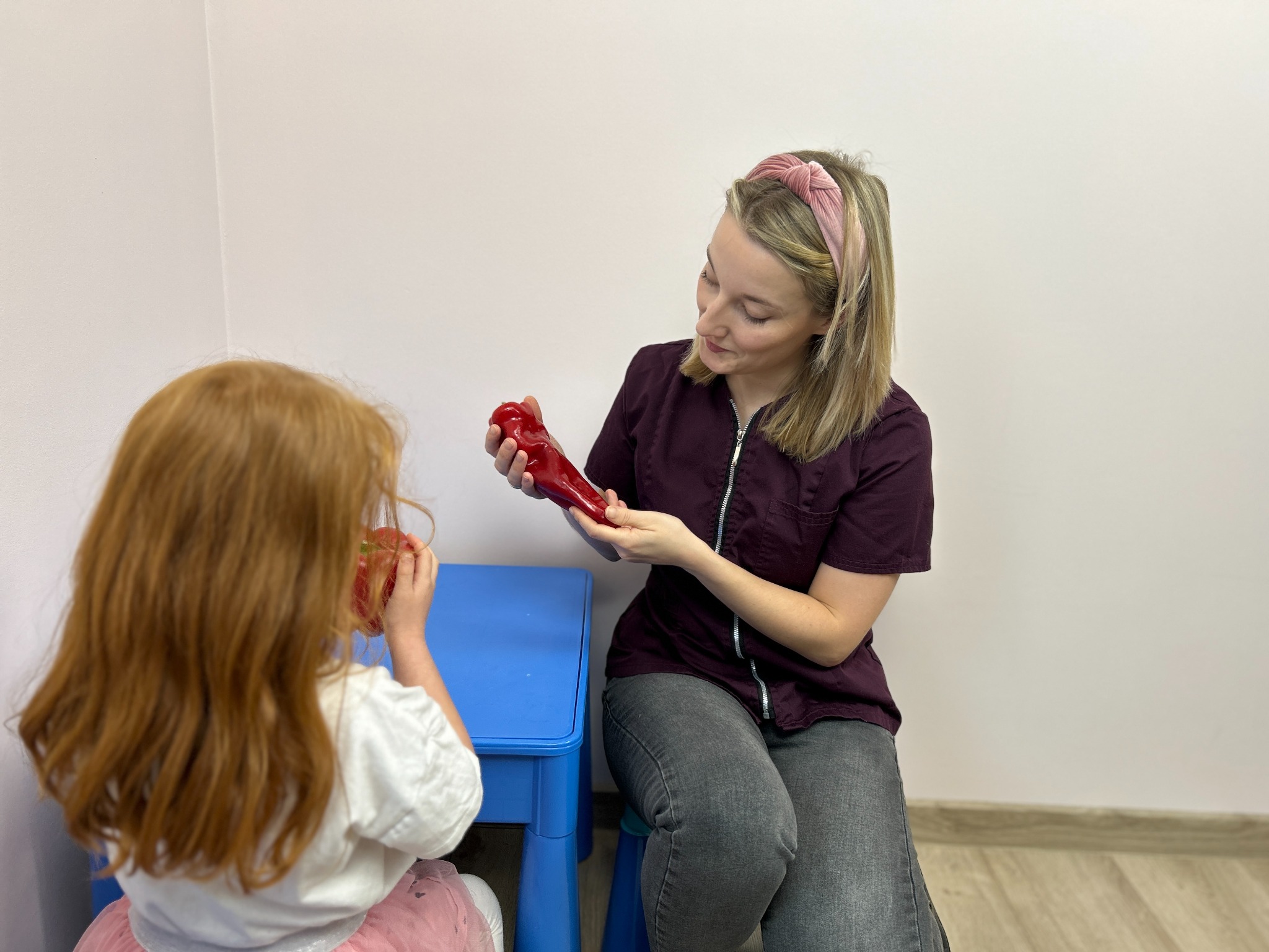 Konsultacja dietetyczna: kobieta w bordowej bluzce prezentuje model papryki dziewczynce przy niebieskim stoliku. Jasne ściany i podłoga.