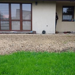 Osoba prywatna - Przygotowany teren pod taras z wysypanym drobnym kamieniem, widoczne fragmenty trawnika i elewacja budynku z oknami.