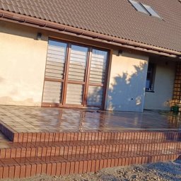 Osoba prywatna - Nowy taras z kostki brukowej w odcieniach brązu, prowadzący do drzwi balkonowych domu jednorodzinnego, w tle widoczne panele solarne na dachu.