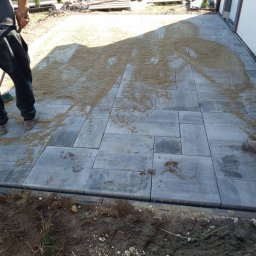 Osoba prywatna - Szare, prostokątne płyty chodnikowe ułożone na zewnątrz budynku, częściowo zasypane piaskiem, z widocznym pracownikiem z miotłą i fragmentem trawnika z tujami w tle.