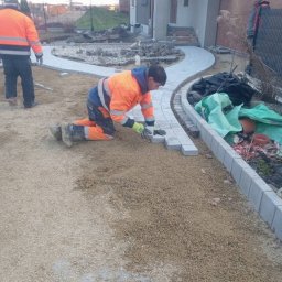 Osoba prywatna - Pracownik w pomarańczowej kurtce klęczy układając szare kostki chodnikowe wzdłuż zakrzywionej ścieżki przed domem, widoczny drugi pracownik w tle.