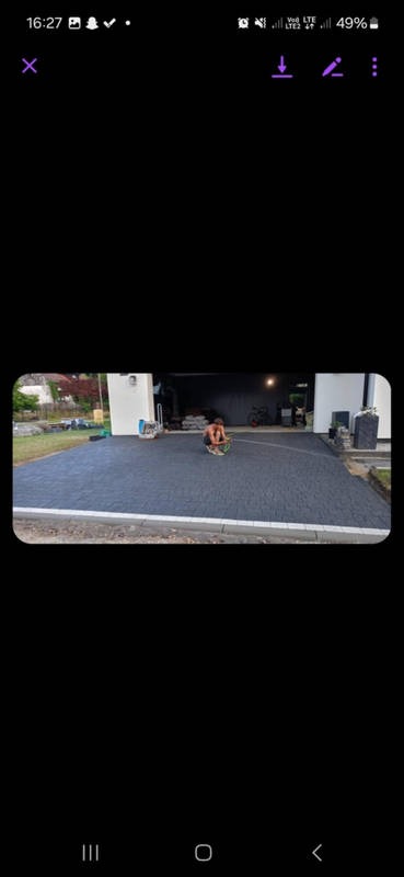 Mężczyzna na kolanach równa świeżo ułożoną, ciemnoszarą nawierzchnię z kostki brukowej przed garażem, z widocznymi narzędziami i workami z materiałem w tle.