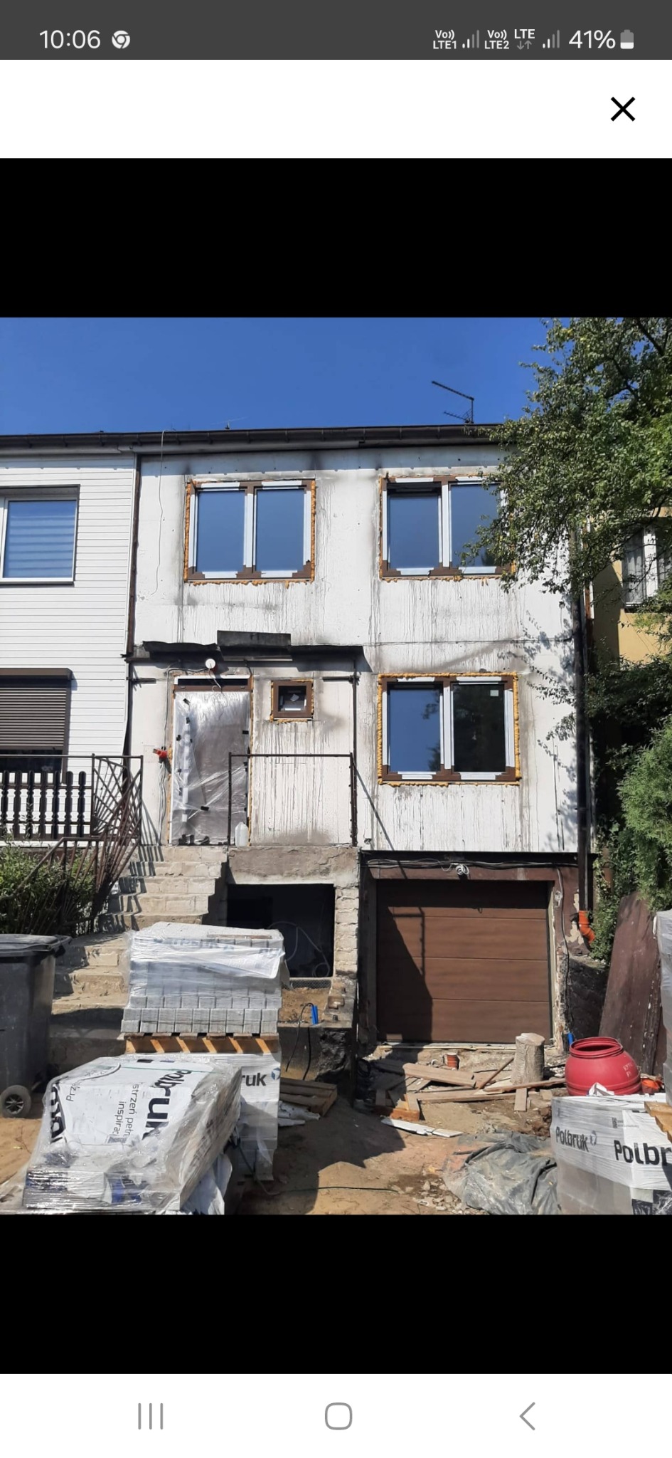 Dom w trakcie remontu elewacji i układania kostki brukowej, widoczne pakiety kostki Polbruk, okna z pianką montażową, ślady po pożarze na elewacji, garaż z brązową bramą.