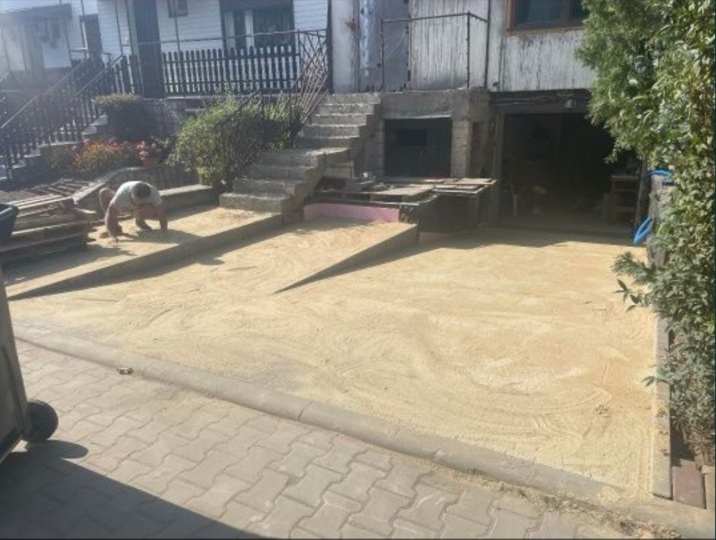 Przygotowanie podłoża pod nawierzchnię: pracownik wyrównuje piasek przed budynkiem z betonowymi schodami i garażem.