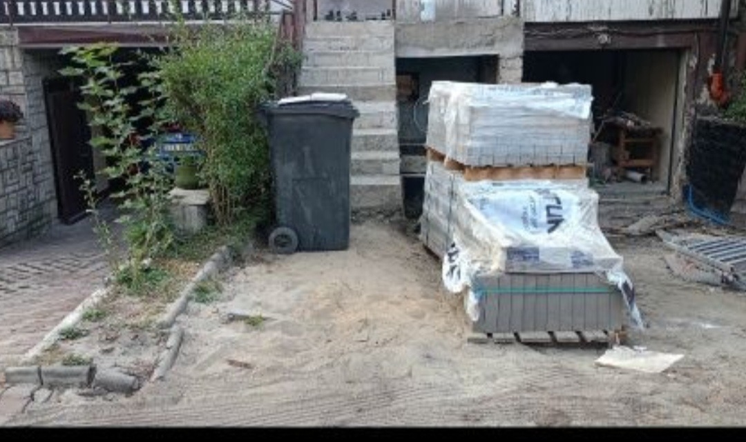 Przygotowany teren pod budowę, palety z kostką brukową zabezpieczone folią, widoczny śmietnik na kółkach i schody wejściowe do budynku.