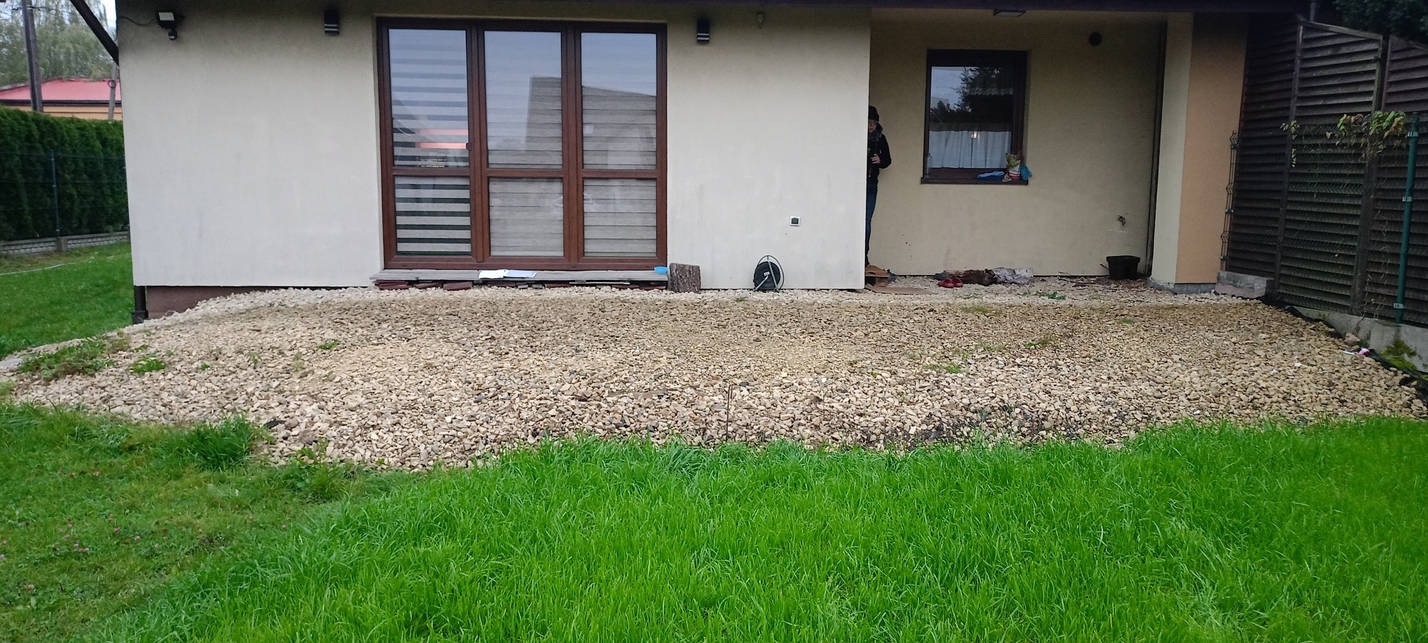 Przygotowany teren pod taras z wysypanym drobnym kamieniem, widoczne fragmenty trawnika i elewacja budynku z oknami.