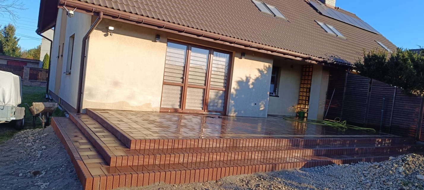 Nowy taras z kostki brukowej w odcieniach brązu, prowadzący do drzwi balkonowych domu jednorodzinnego, w tle widoczne panele solarne na dachu.