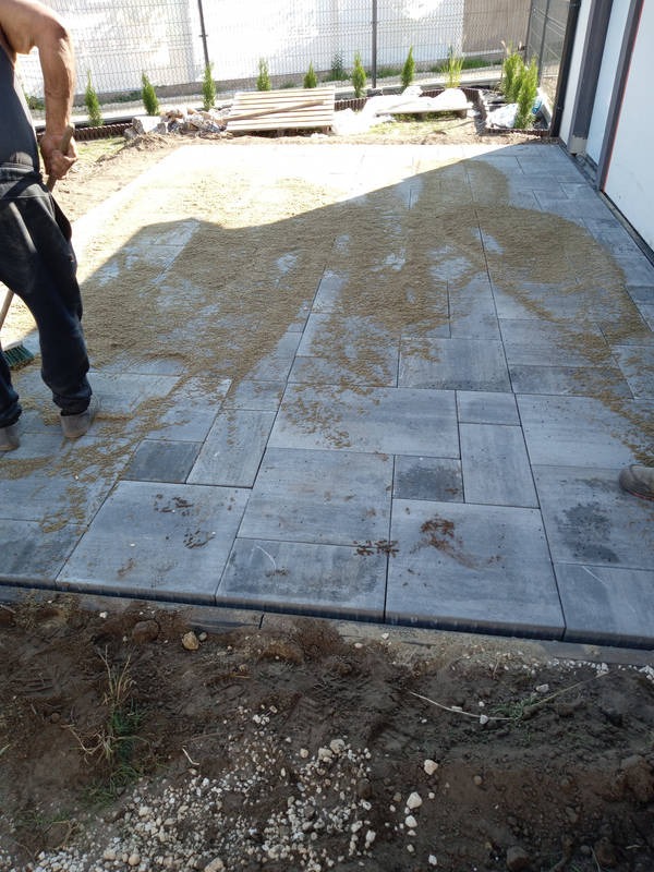 Szare, prostokątne płyty chodnikowe ułożone na zewnątrz budynku, częściowo zasypane piaskiem, z widocznym pracownikiem z miotłą i fragmentem trawnika z tujami w tle.