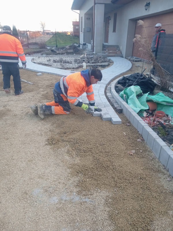 Pracownik w pomarańczowej kurtce klęczy układając szare kostki chodnikowe wzdłuż zakrzywionej ścieżki przed domem, widoczny drugi pracownik w tle.