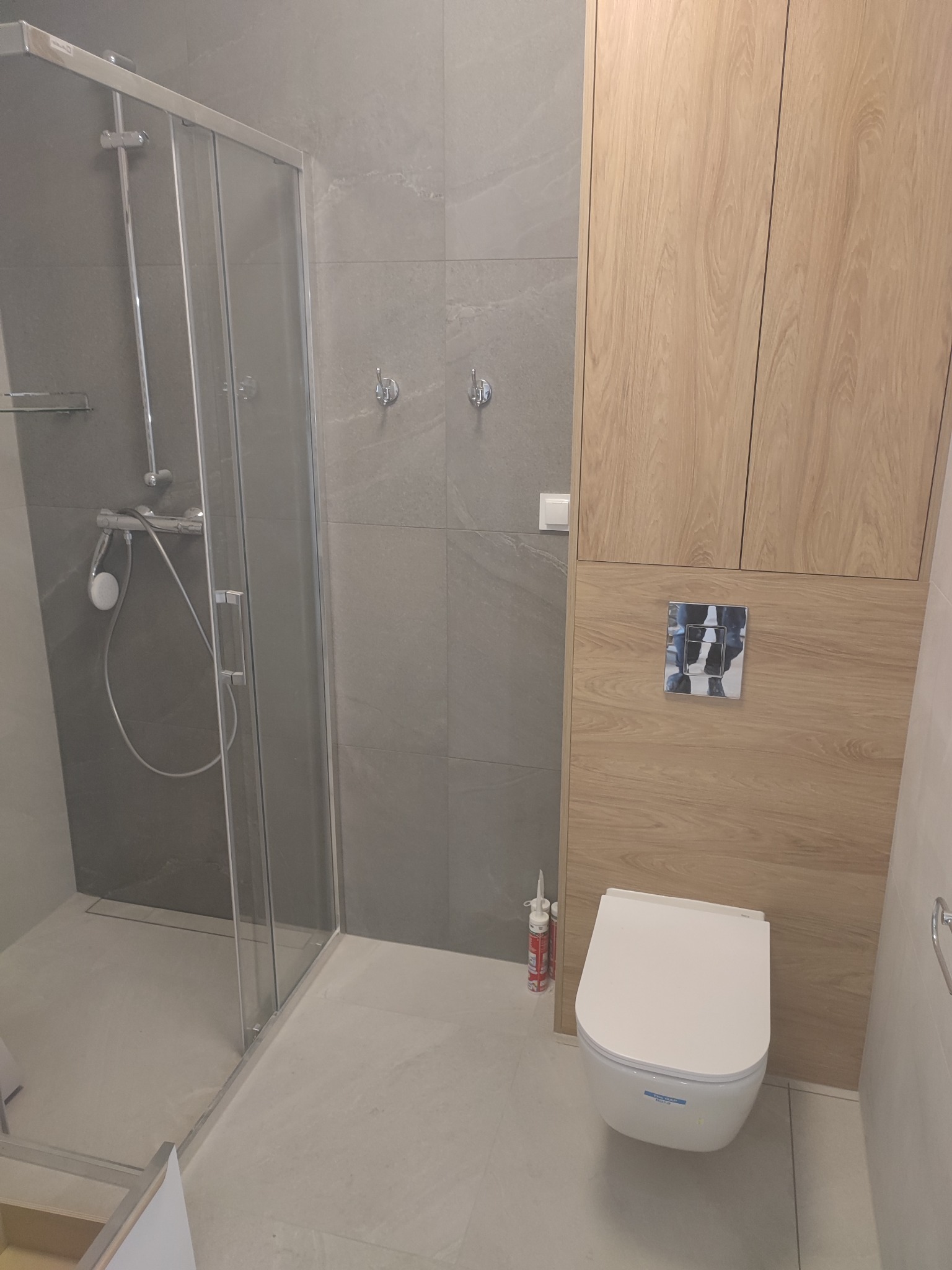 Nowoczesna łazienka z kabiną prysznicową typu walk-in, wiszącą toaletą i zabudową meblową z drewnopodobnym wykończeniem, szare płytki na ścianach i podłodze, widoczna tuba silikonu sanitarny przy...