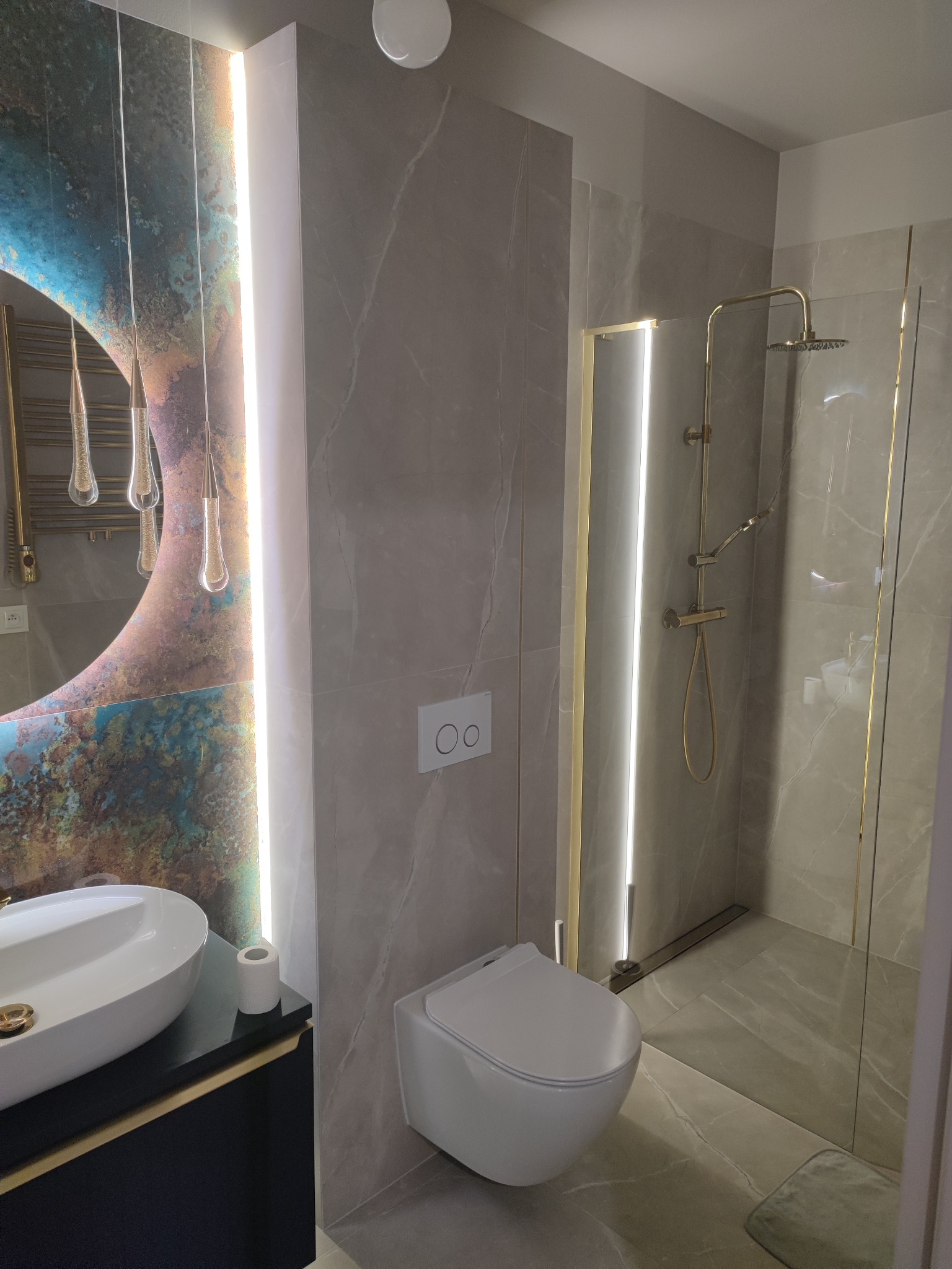 Elegancka, wykończona łazienka w Pasłęku z szarymi płytkami imitującymi marmur, okrągłym lustrem z podświetleniem LED i złotymi akcentami w kabinie prysznicowej oraz armaturze.