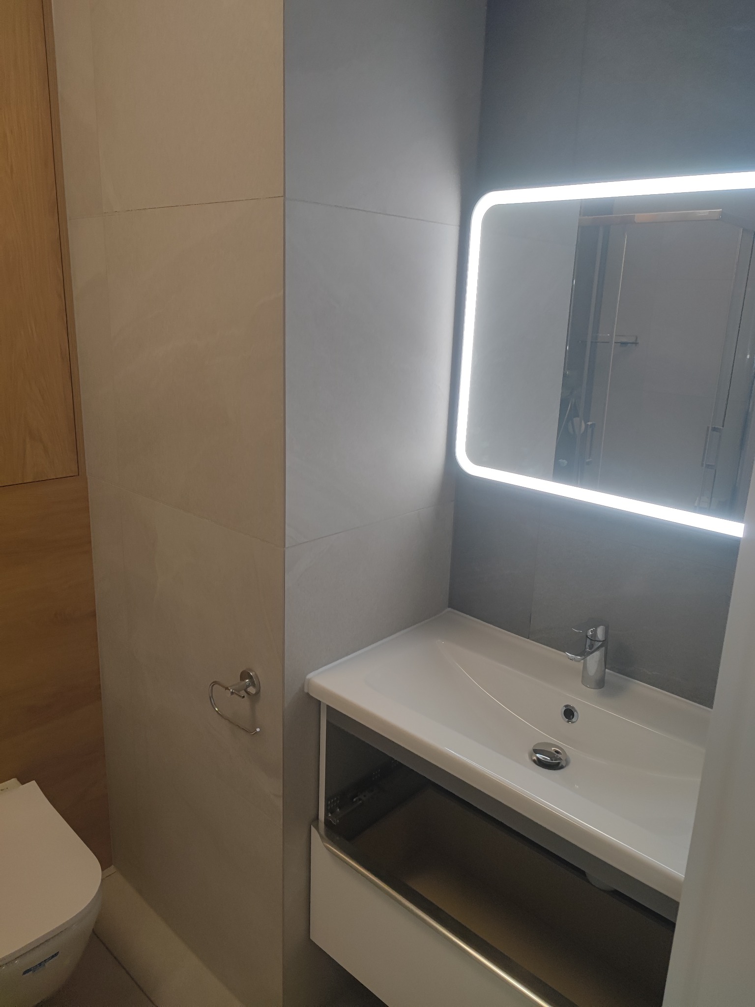 Nowoczesna łazienka z umywalką w szafce z otwartą szufladą, podświetlanym lustrem LED i beżowymi płytkami imitującymi marmur.