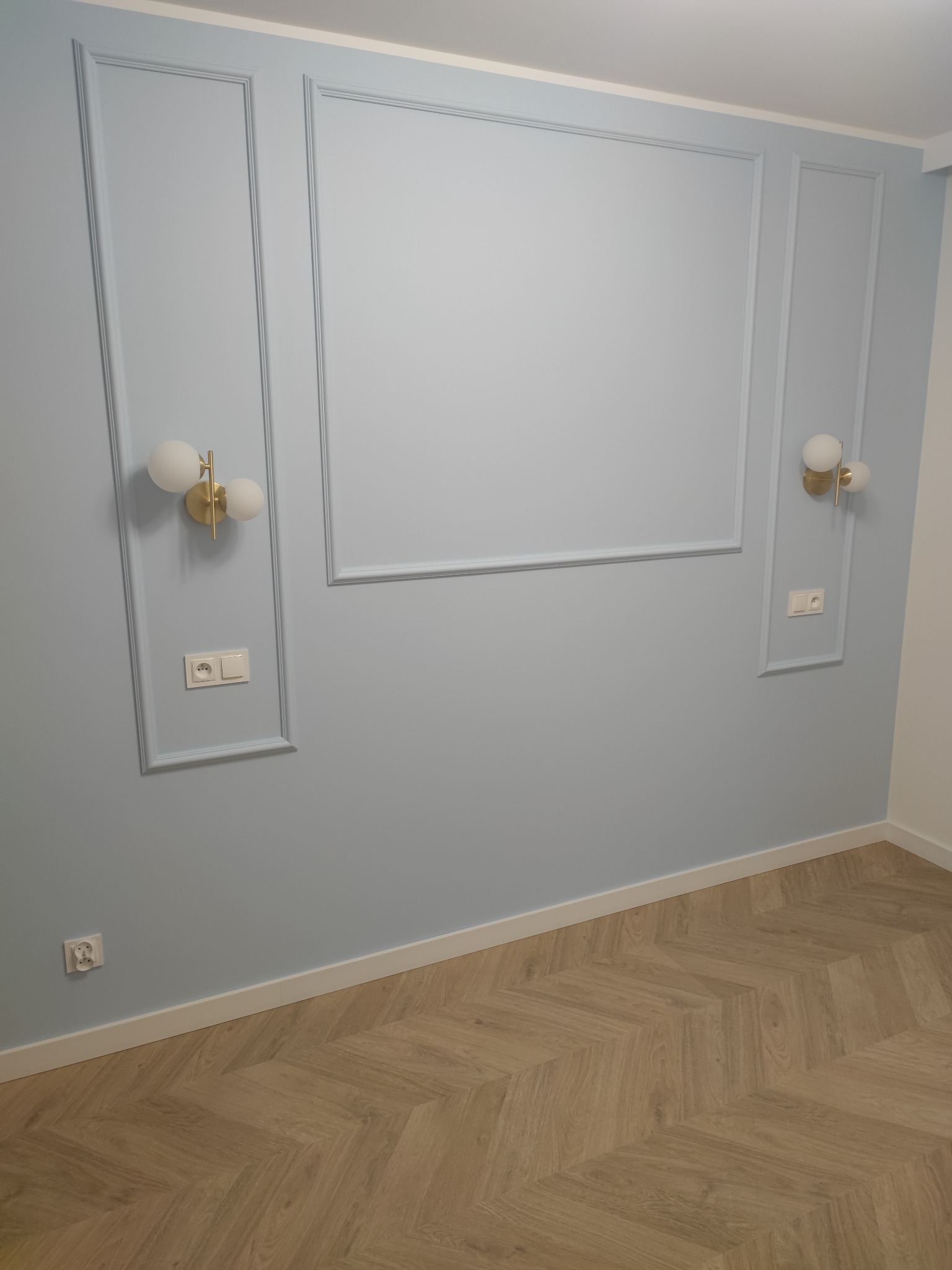 Ściana w kolorze gołębiej szarości z dekoracyjnymi ramkami, złotymi kinkietami z białymi kulami i parkietem ułożonym w jodełkę