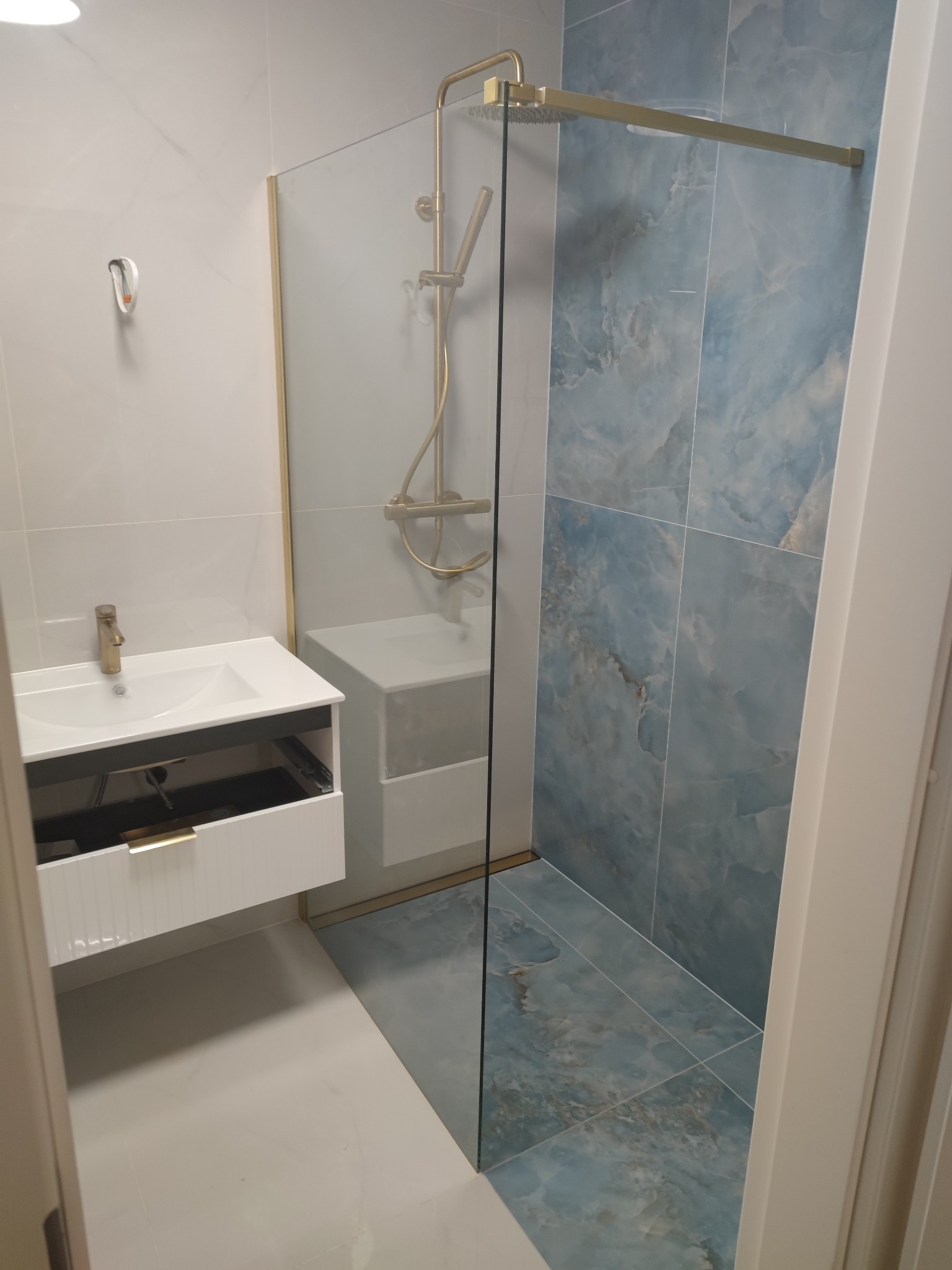 Nowoczesna łazienka z kabiną prysznicową typu walk-in, umywalką nablatową z szafką z otwartą szufladą i niebieskimi płytkami imitującymi marmur na ścianach.