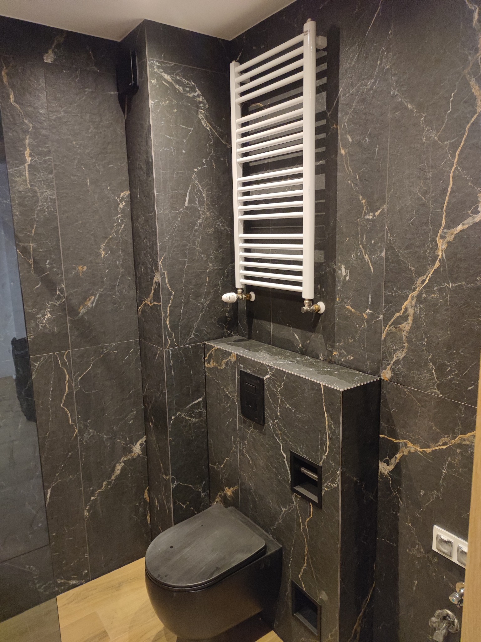 Nowoczesna łazienka wykończona ciemnymi płytkami imitującymi marmur ze złotymi żyłami, wisząca toaleta w kolorze grafitowym, biały grzejnik drabinkowy na ścianie, drewnopodobna podłoga.