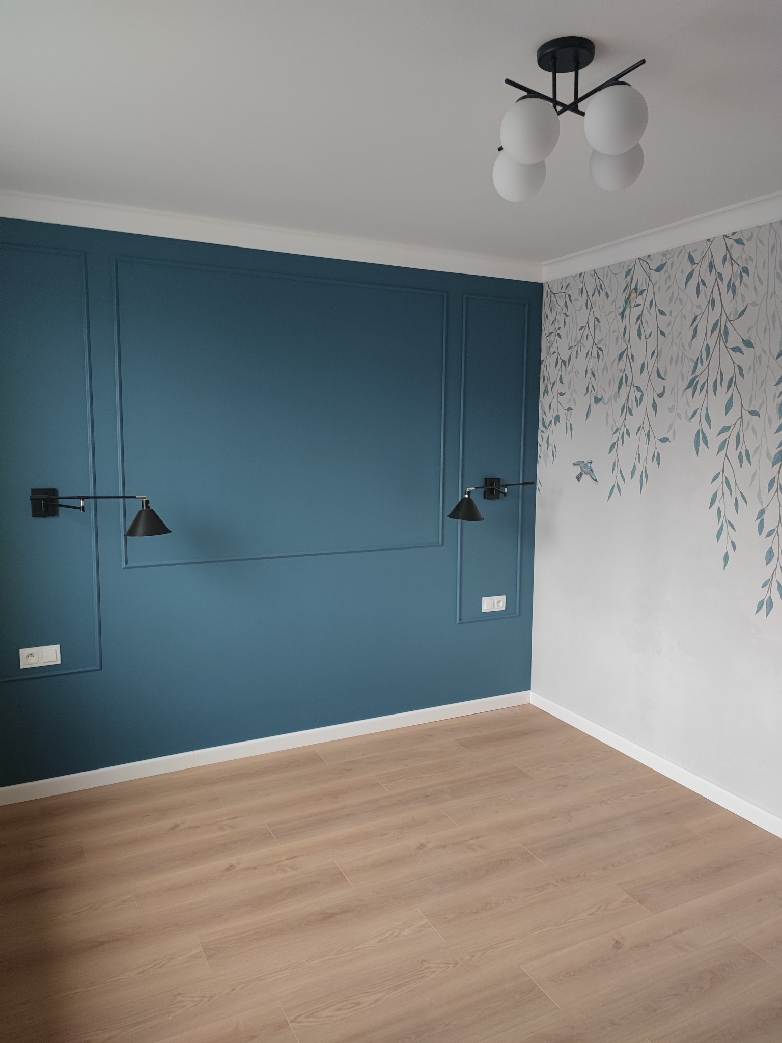 Pusty pokój po remoncie z panelami podłogowymi, ścianą w kolorze morskim z dekoracyjnymi listwami, ścianą z motywem liści i lampami ściennymi oraz sufitową lampą kulistą.