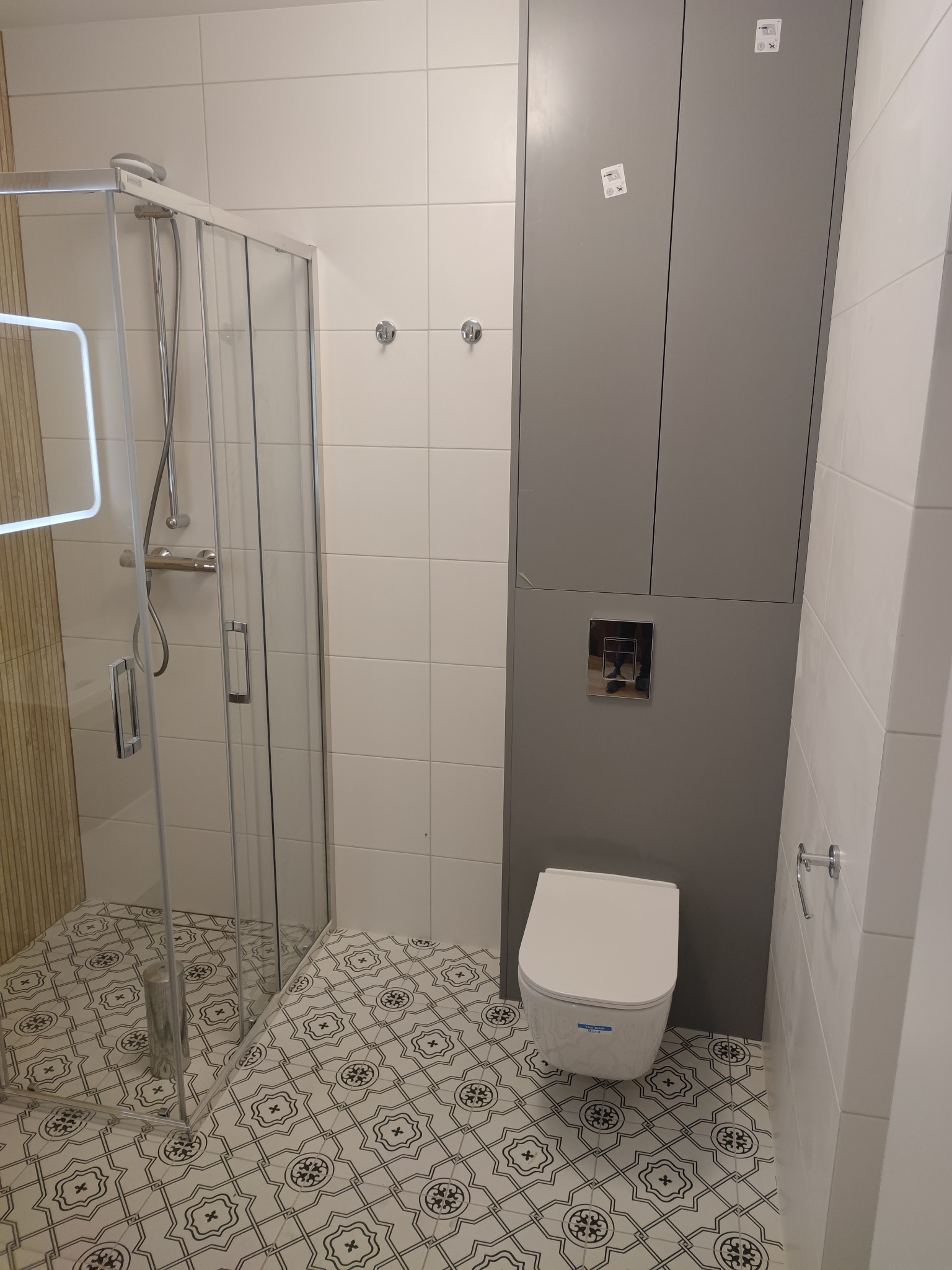 Nowoczesna łazienka z kabiną prysznicową ze szklanymi drzwiami, podwieszaną toaletą i szarą szafką zabudowaną, wzorzysta podłoga w biało-czarne ornamenty.