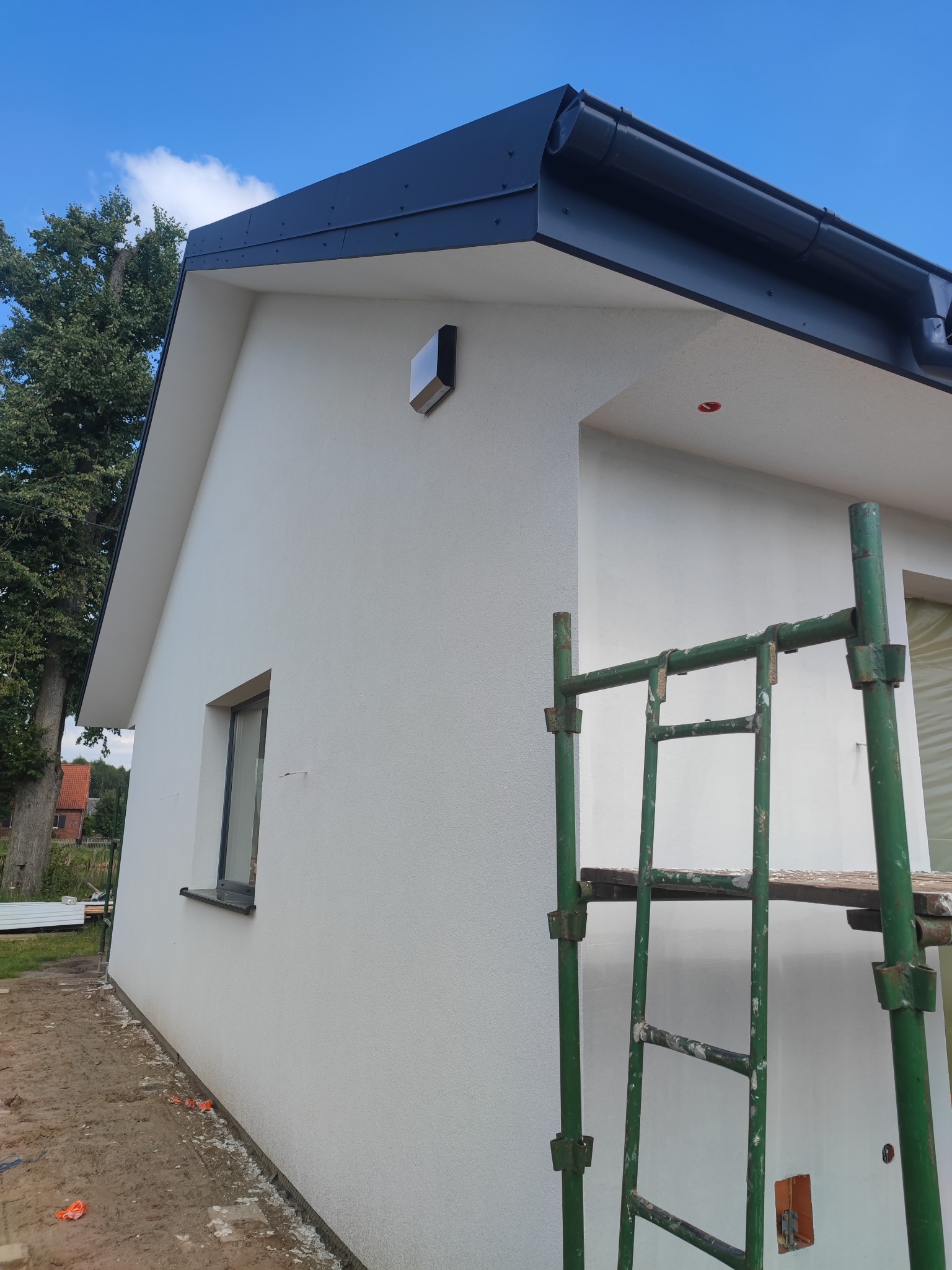 Świeżo otynkowana elewacja domu jednorodzinnego z widocznym rusztowaniem malarskim, czarnym dachem i fragmentem zielonego drzewa w tle.