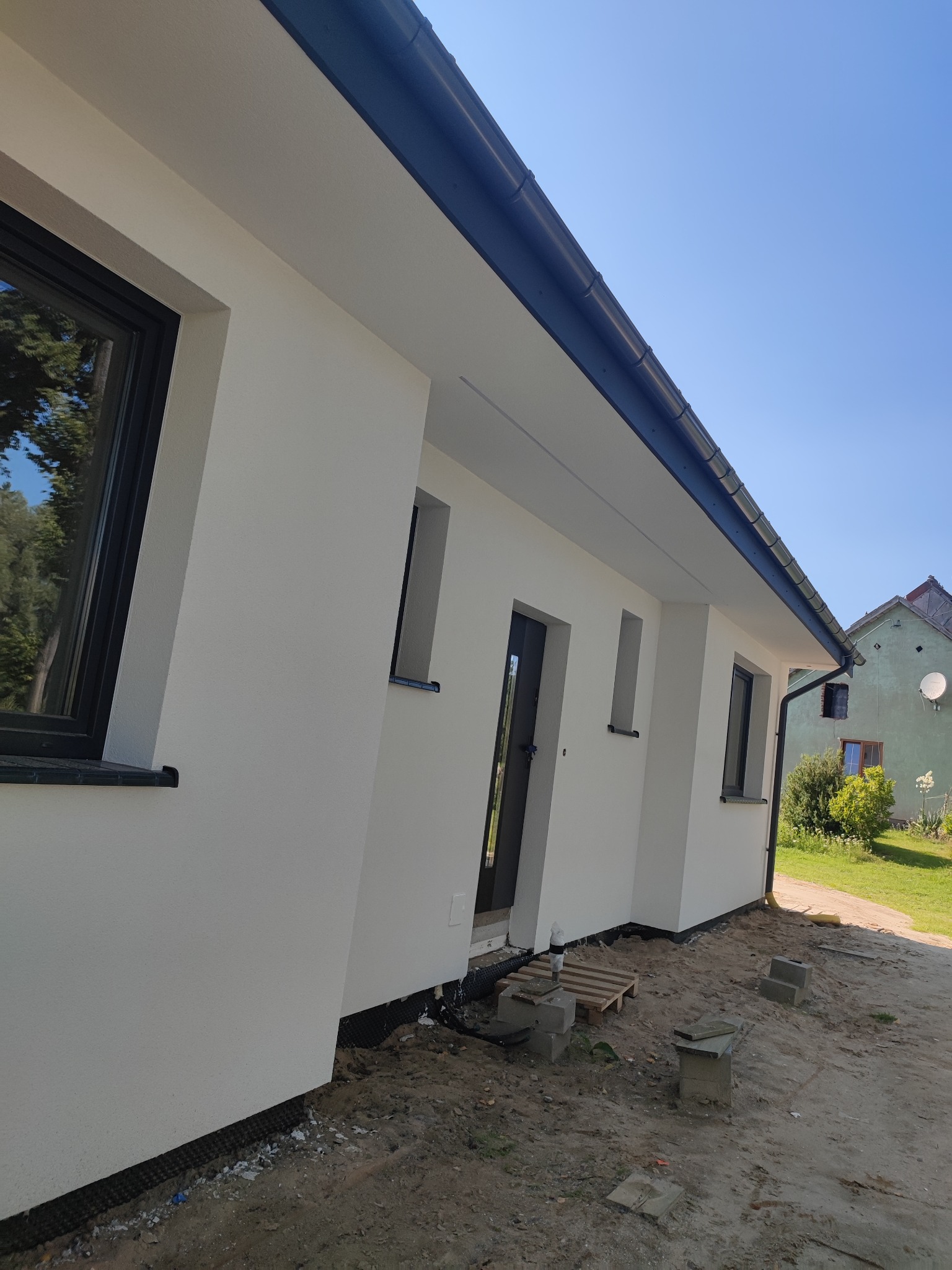 Świeżo otynkowany dom jednorodzinny z czarnymi oknami i rynnami, widok z zewnątrz, teren wokół domu jeszcze niezagospodarowany, widoczne bloczki betonowe i paleta.