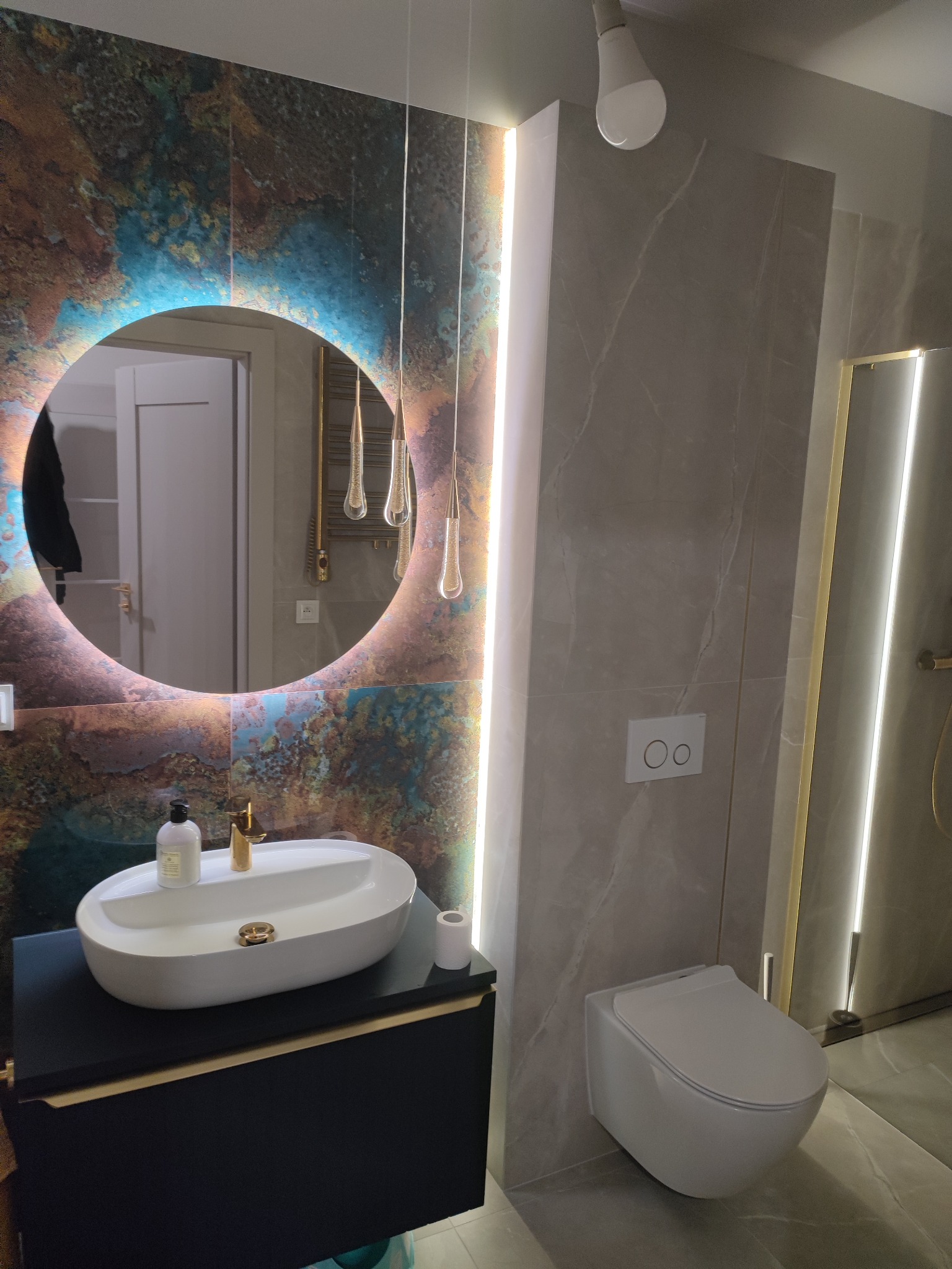 Nowoczesna łazienka z okrągłym lustrem podświetlanym LED, umywalką nablatową na czarnej szafce, wiszącą toaletą i płytkami imitującymi marmur.