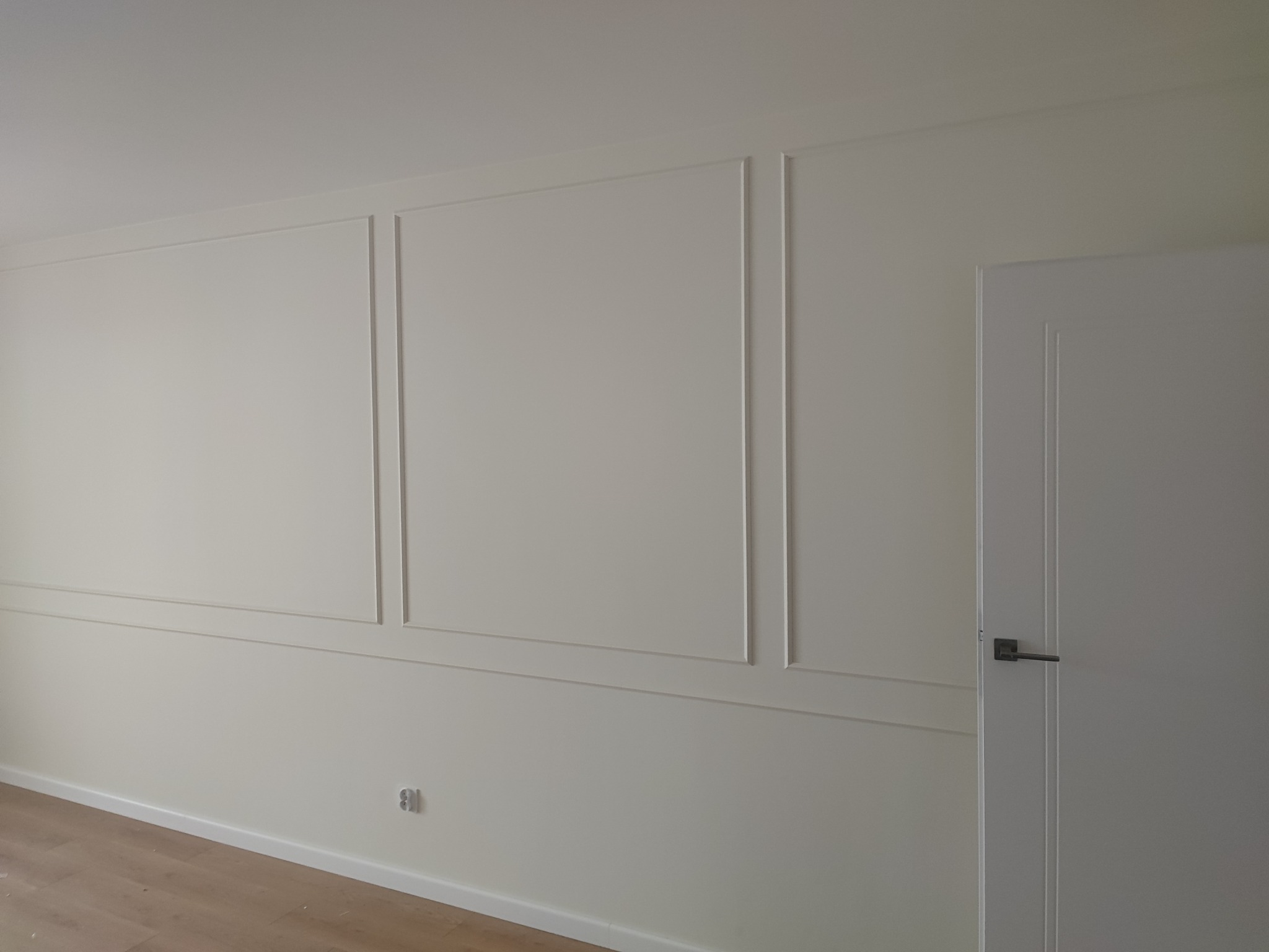 Wykończona ściana z dekoracyjnymi listwami w kolorze kremowym, widoczny fragment podłogi z paneli i białe drzwi z czarną klamką.
