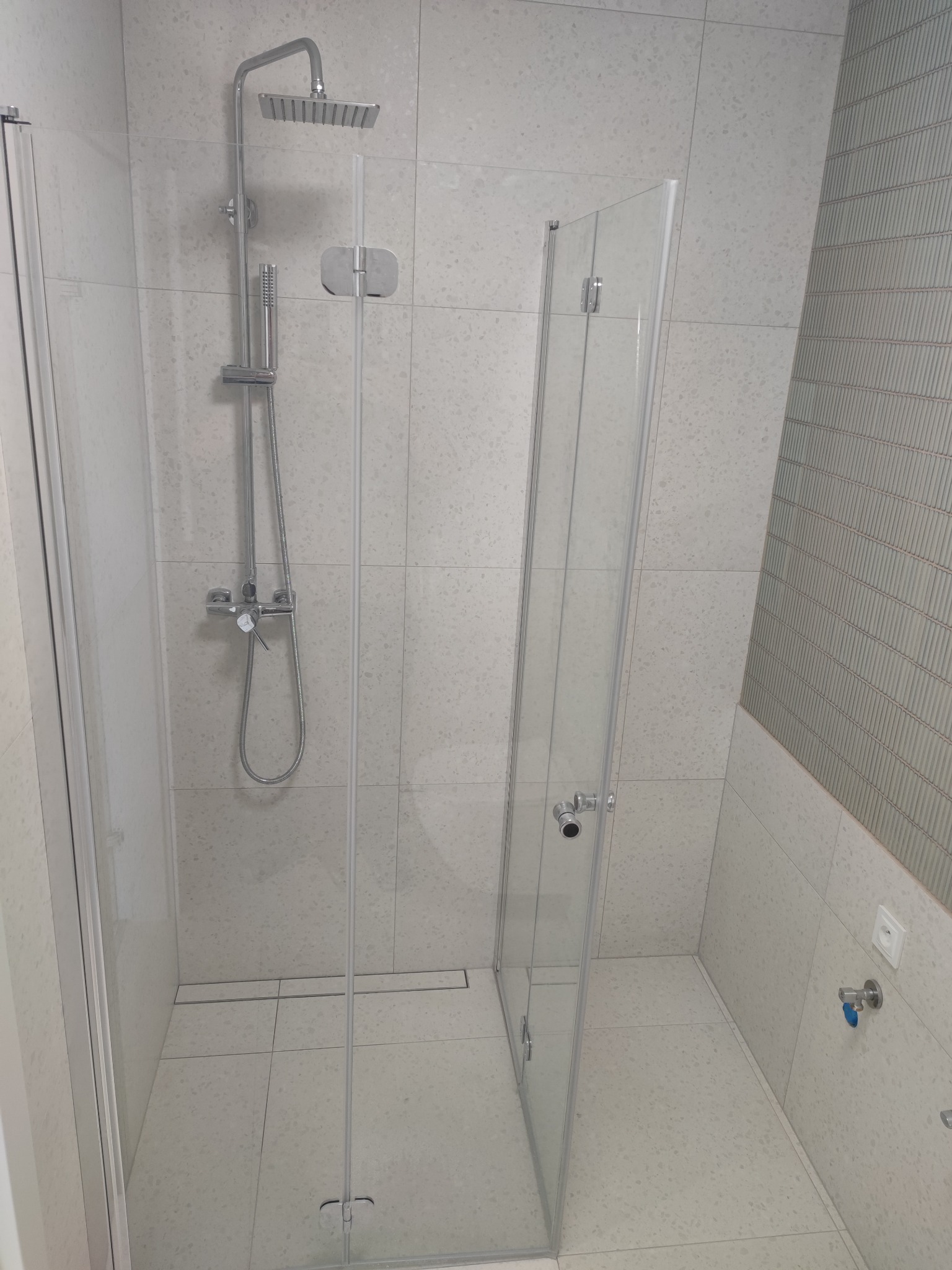 Nowoczesna kabina prysznicowa z deszczownicą i szklanymi drzwiami, wykończona jasnymi płytkami z delikatnym wzorem, widoczny odpływ liniowy w podłodze.