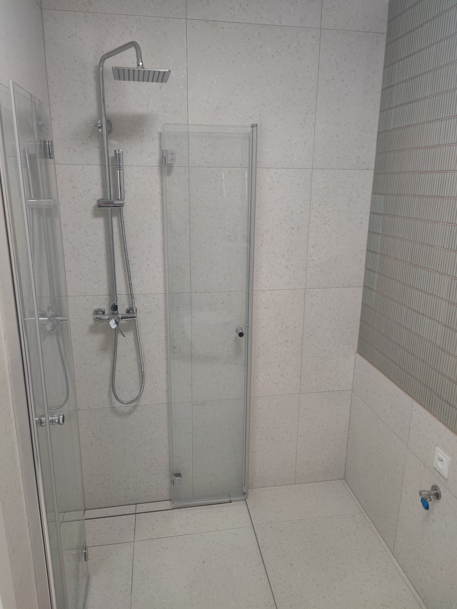 Nowoczesna kabina prysznicowa z deszczownicą i szklanymi drzwiami, wykończona płytkami w odcieniach bieli i szarości, z akcentem dekoracyjnej ściany z pionowymi żłobieniami.