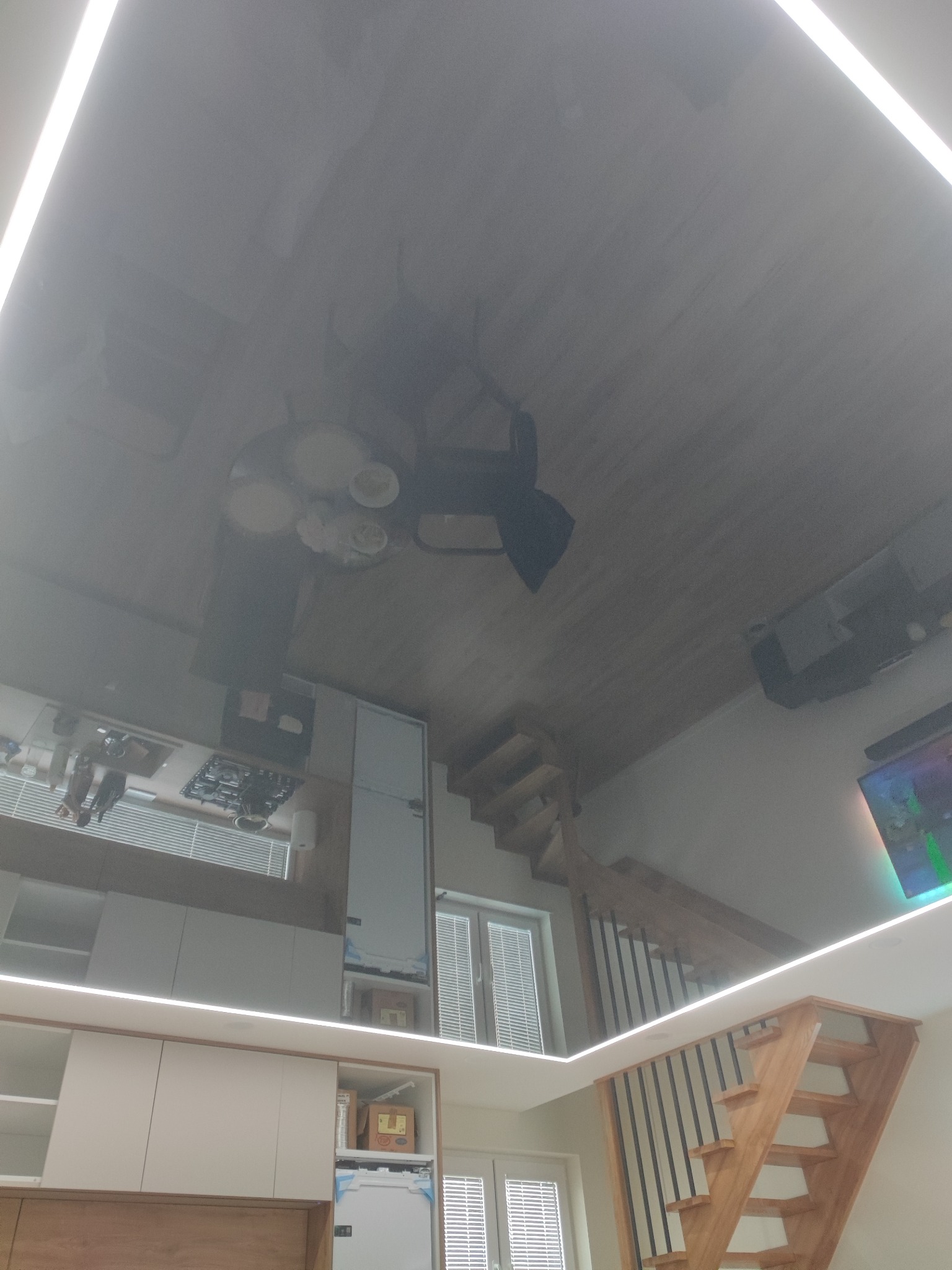 Błyszczący sufit napinany w nowoczesnym mieszkaniu odbija wyposażenie: drewniane schody, białe meble kuchenne i stół z krzesłami.