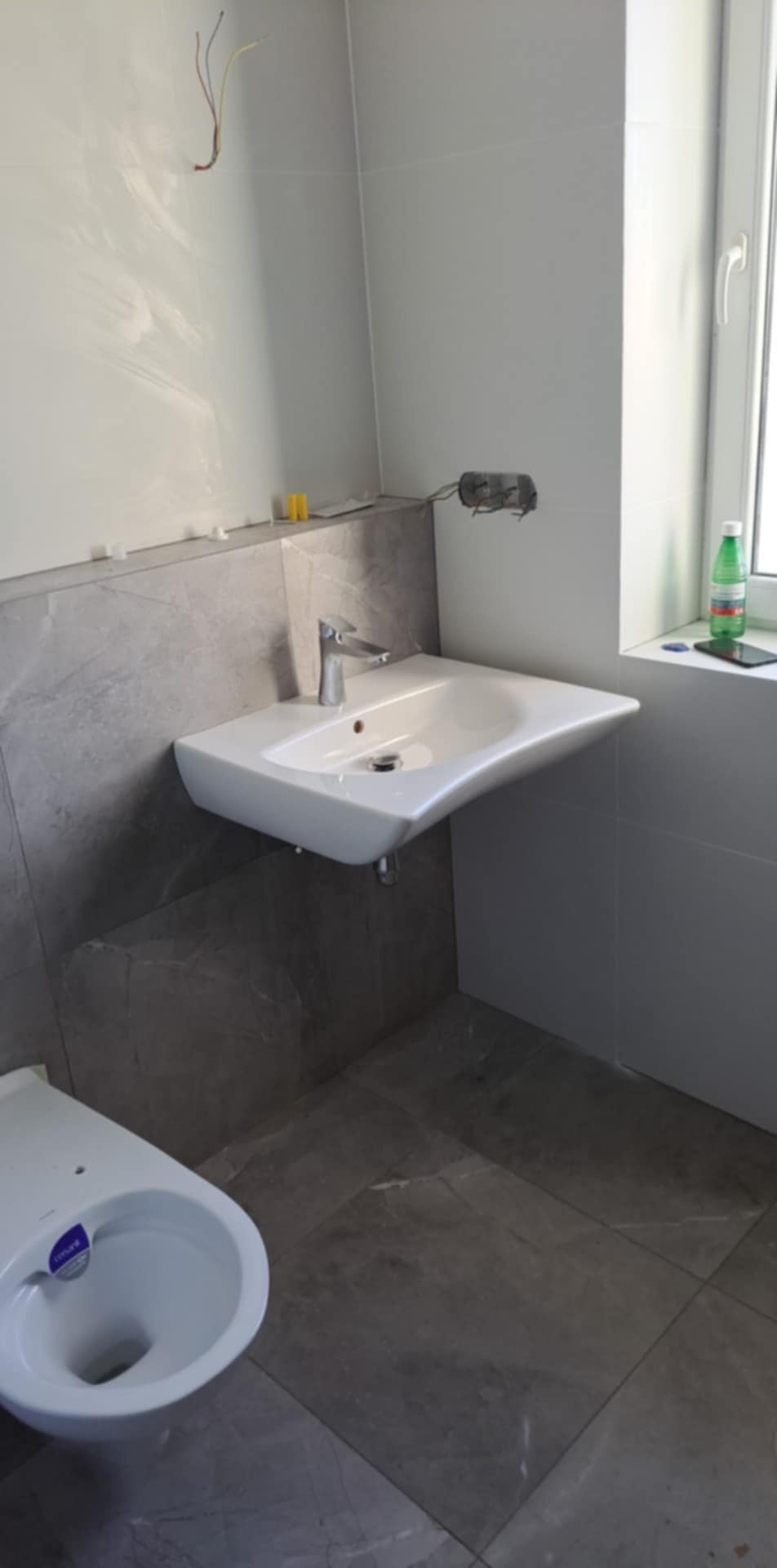 Umywalka podwieszana z baterią zamontowana na ścianie wyłożonej szarymi płytkami imitującymi marmur, widoczna toaleta i wystające przewody elektryczne.