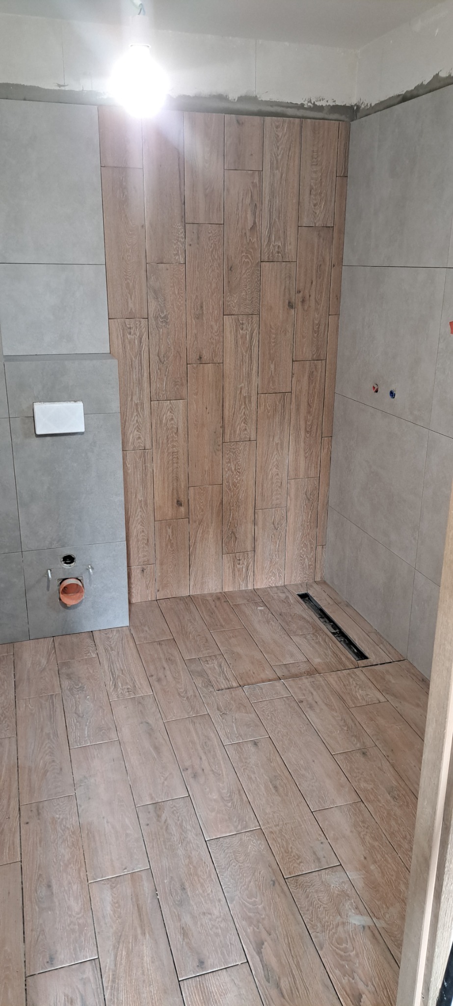Wnętrze łazienki w trakcie układania płytek imitujących drewno na ścianie prysznica i podłodze, z widocznym odpływem liniowym w podłodze oraz szarą płytką na pozostałych ścianach, z przygotowanym...