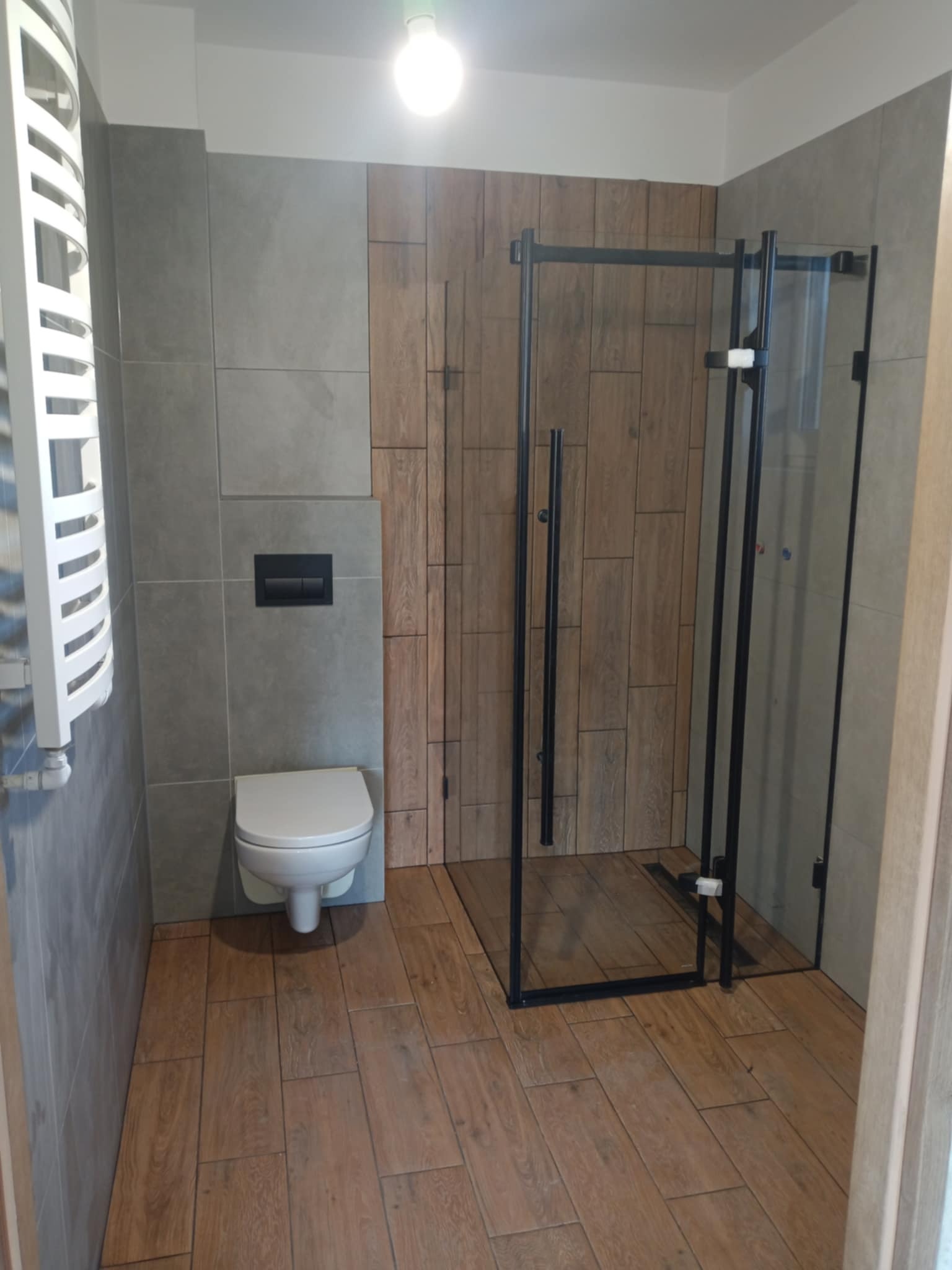 Nowoczesna łazienka z kabiną prysznicową typu walk-in, drewnopodobnymi i szarymi płytkami. Uwagę zwraca grzejnik drabinkowy i podwieszana toaleta.