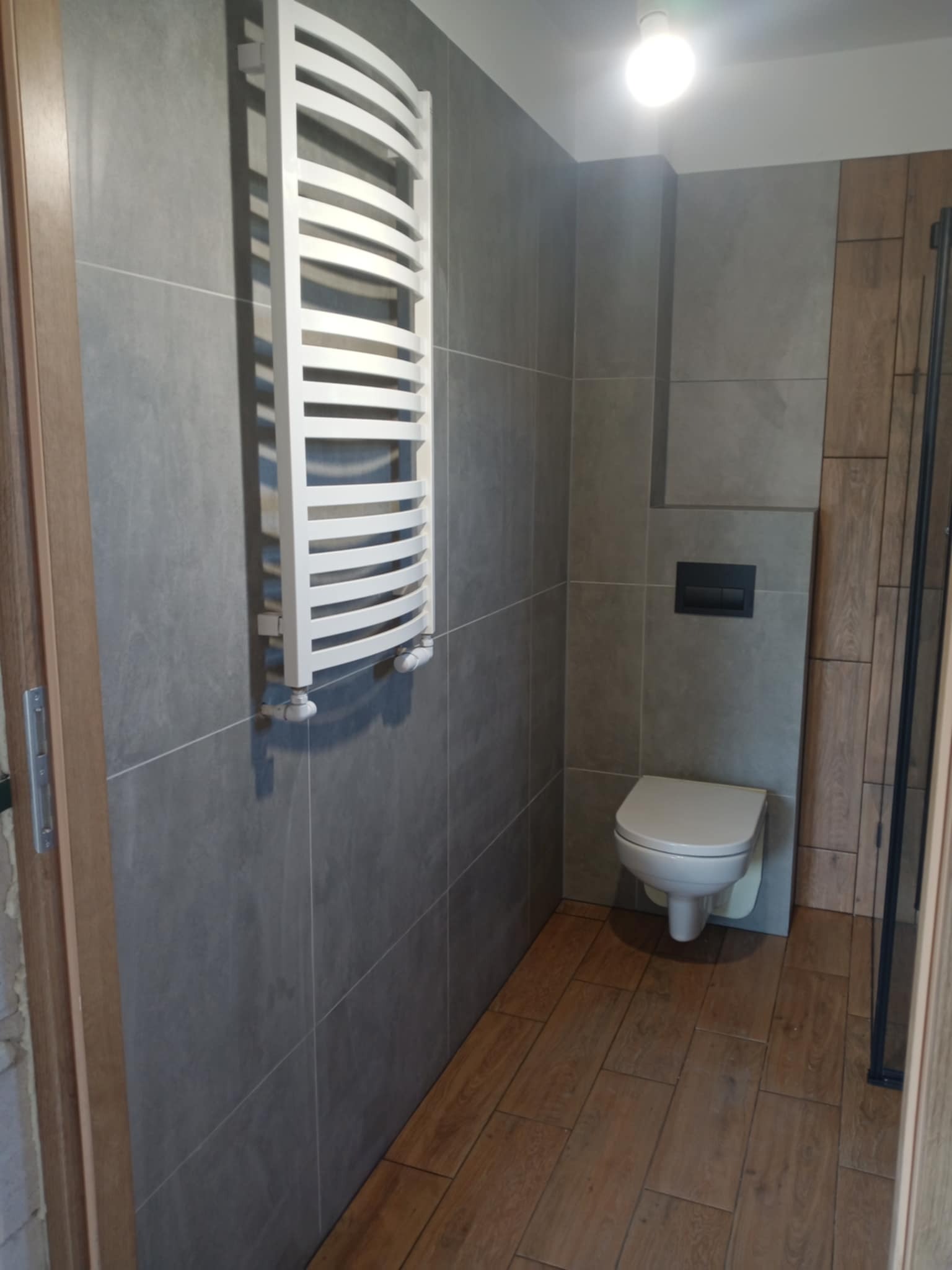 Nowoczesna łazienka z szarymi płytkami, drewnopodobną podłogą i białym grzejnikiem drabinkowym. Uwagę zwraca minimalistyczna toaleta podwieszana.