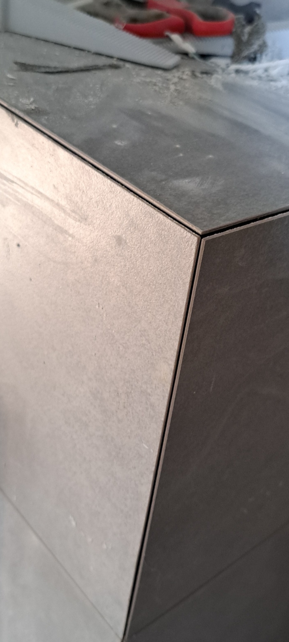 Precyzyjnie ułożone, szare płytki ceramiczne na rogu ściany, z widocznymi narzędziami glazurniczymi w tle. Detal wykończenia, wąska spoina, ostre krawędzie.