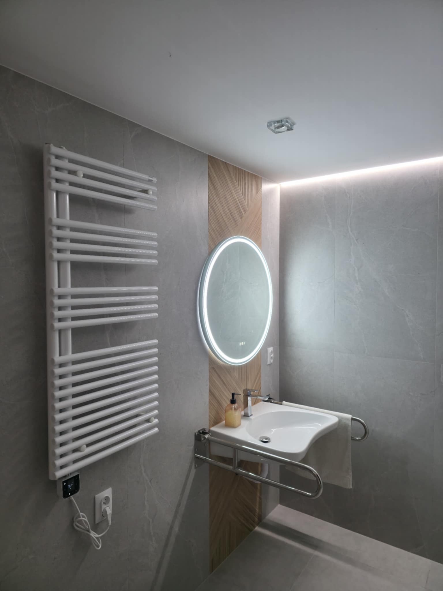 Nowoczesna łazienka z szarymi płytkami, podświetlanym lustrem, grzejnikiem drabinkowym i umywalką z uchwytami. Drewniane panele dekoracyjne dodają ciepła.