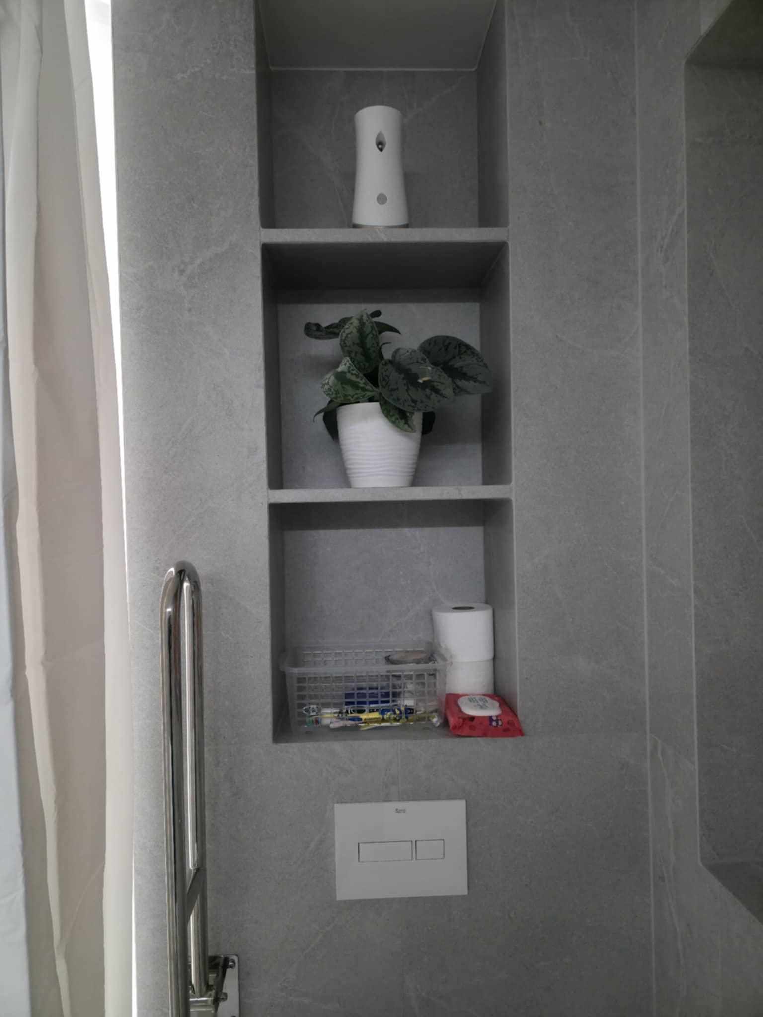 Wnęka w łazience wyłożona szarą płytką imitującą kamień. W półkach dekoracje: roślina w doniczce, odświeżacz powietrza, koszyk z drobiazgami i papier toaletowy. Widoczny uchwyt dla niepełnosprawnych.