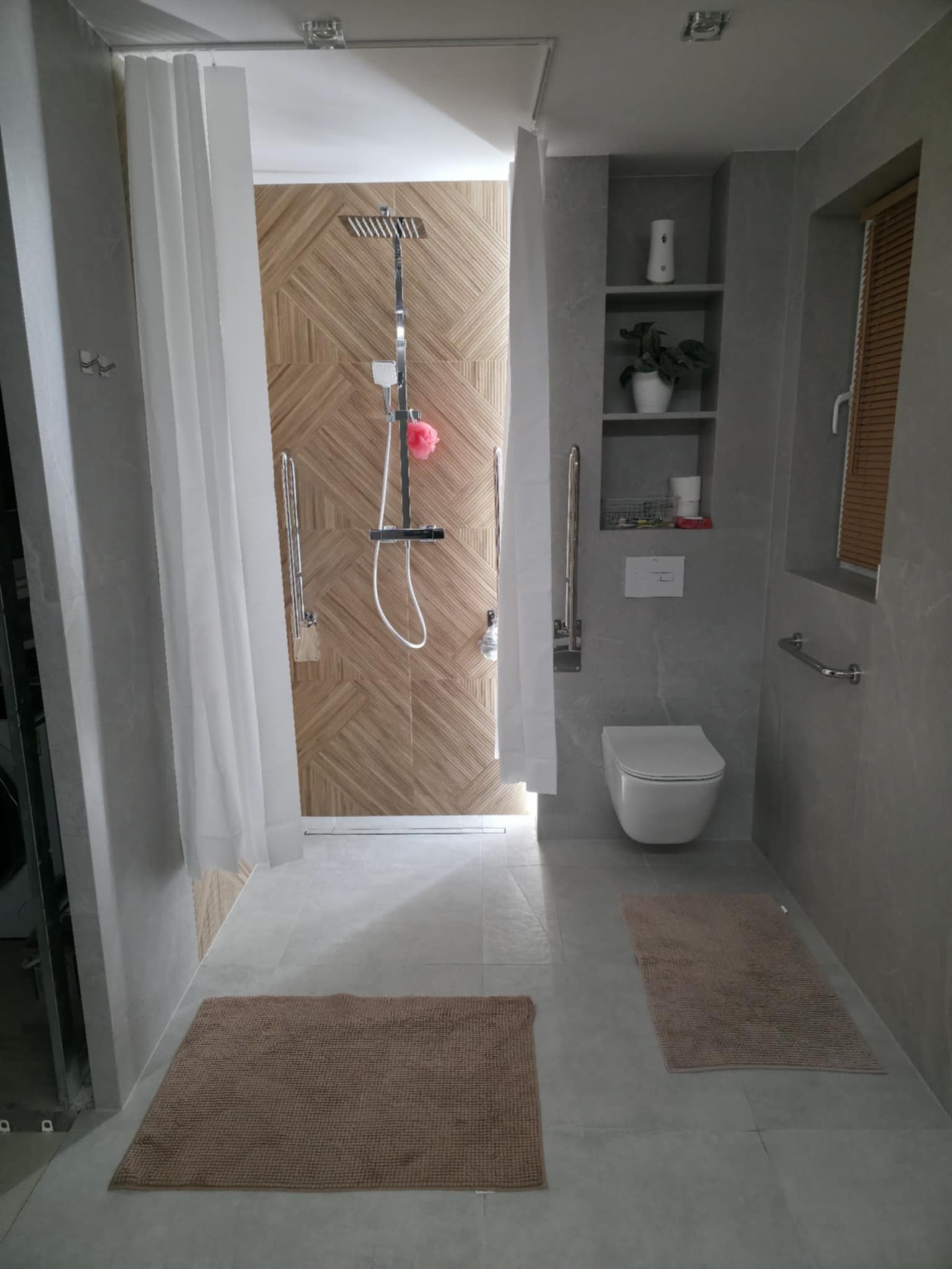 Nowoczesna łazienka z kabiną prysznicową typu walk-in, wykończona płytkami imitującymi drewno i szarą ceramiką. Widoczne detale: deszczownica, półki wnękowe i dywaniki.