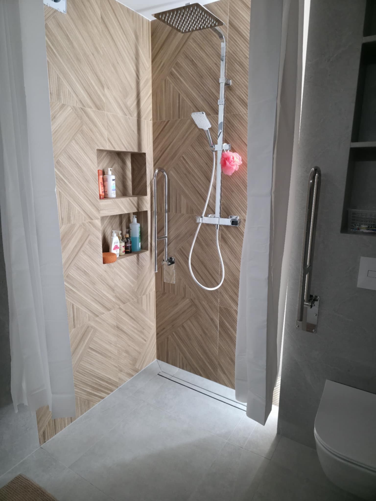 Nowoczesna kabina prysznicowa z drewnopodobnymi płytkami, wnękami na kosmetyki i szklanym odpływem liniowym. Uchwyt dla niepełnosprawnych.