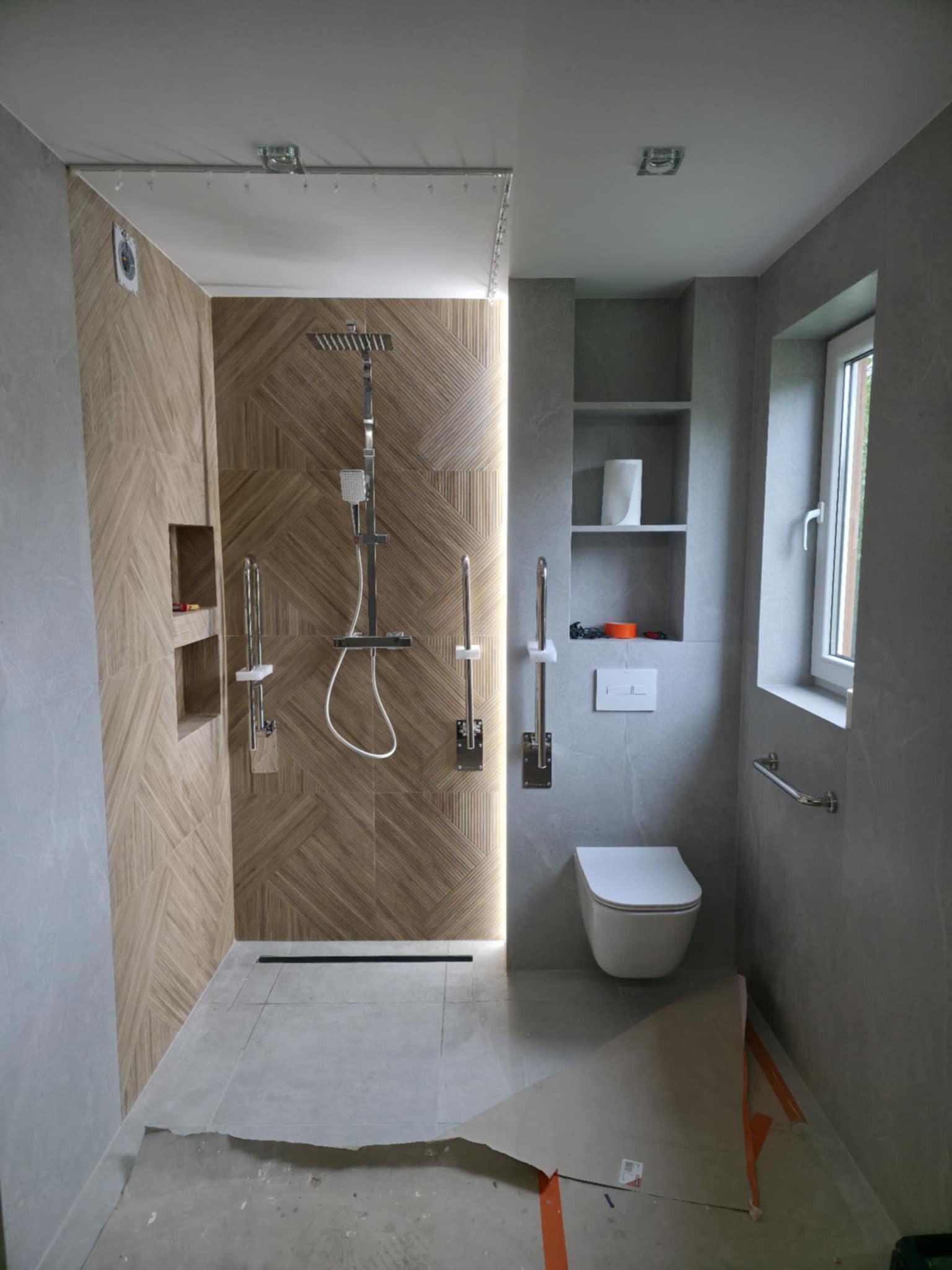 Nowoczesna łazienka z kabiną typu walk-in, wykończona szarą i drewnopodobną płytką. Widoczne detale: deszczownica, uchwyty dla niepełnosprawnych, wnęki w ścianie.
