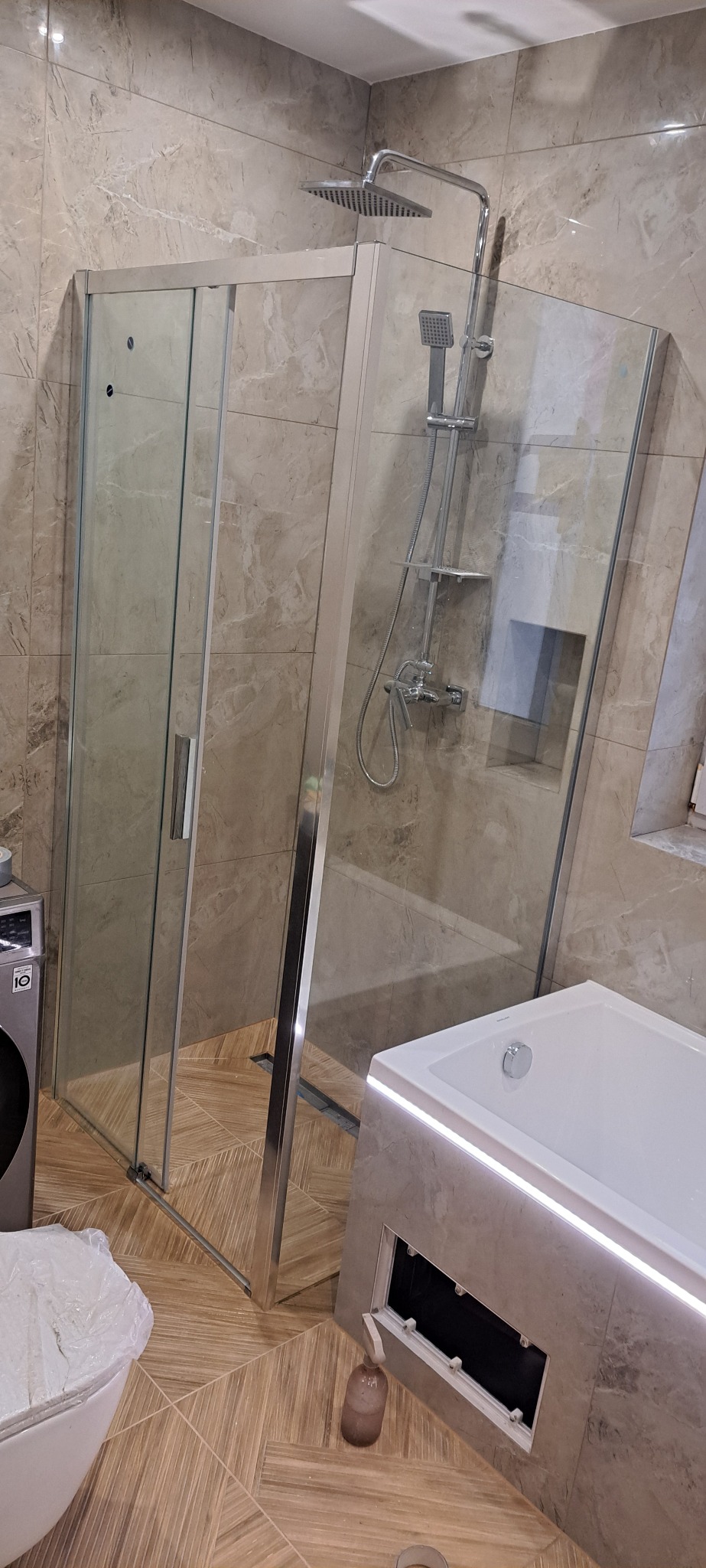 Nowoczesna kabina prysznicowa ze szklanymi drzwiami i deszczownicą, obok wanna z podświetleniem LED. Beżowe płytki imitujące marmur na ścianach i podłodze.